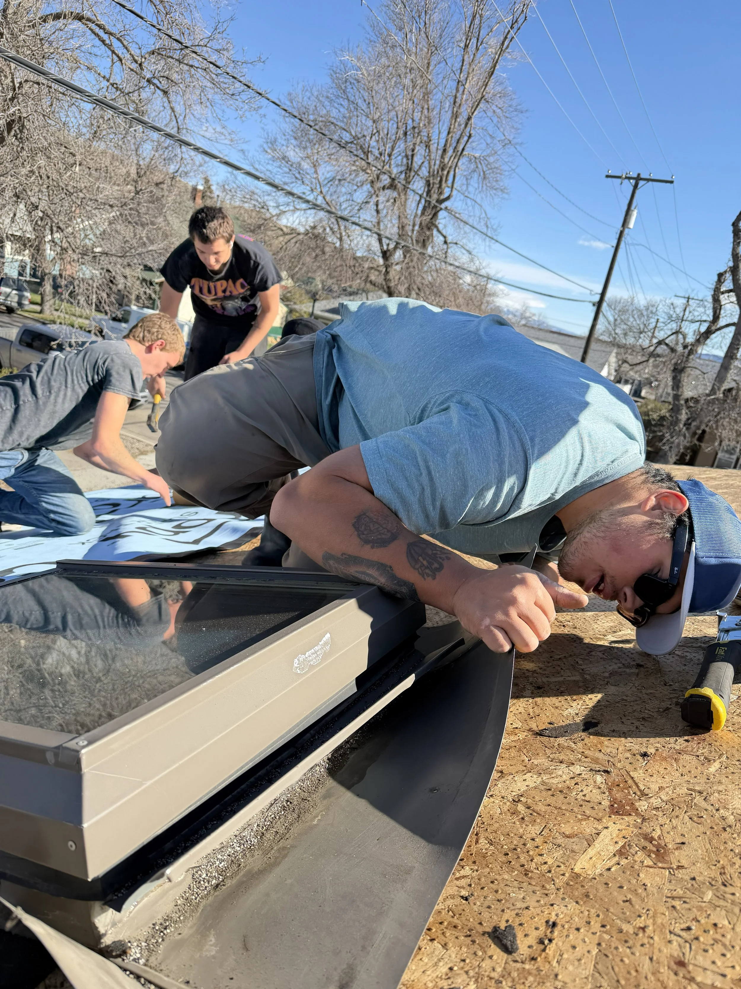 Group of people working on a construction project outdoors on a clear, sunny day. They are focusing on a large piece of equipment or machinery, with some using tools. In the background, there are trees, power lines, and residential houses.