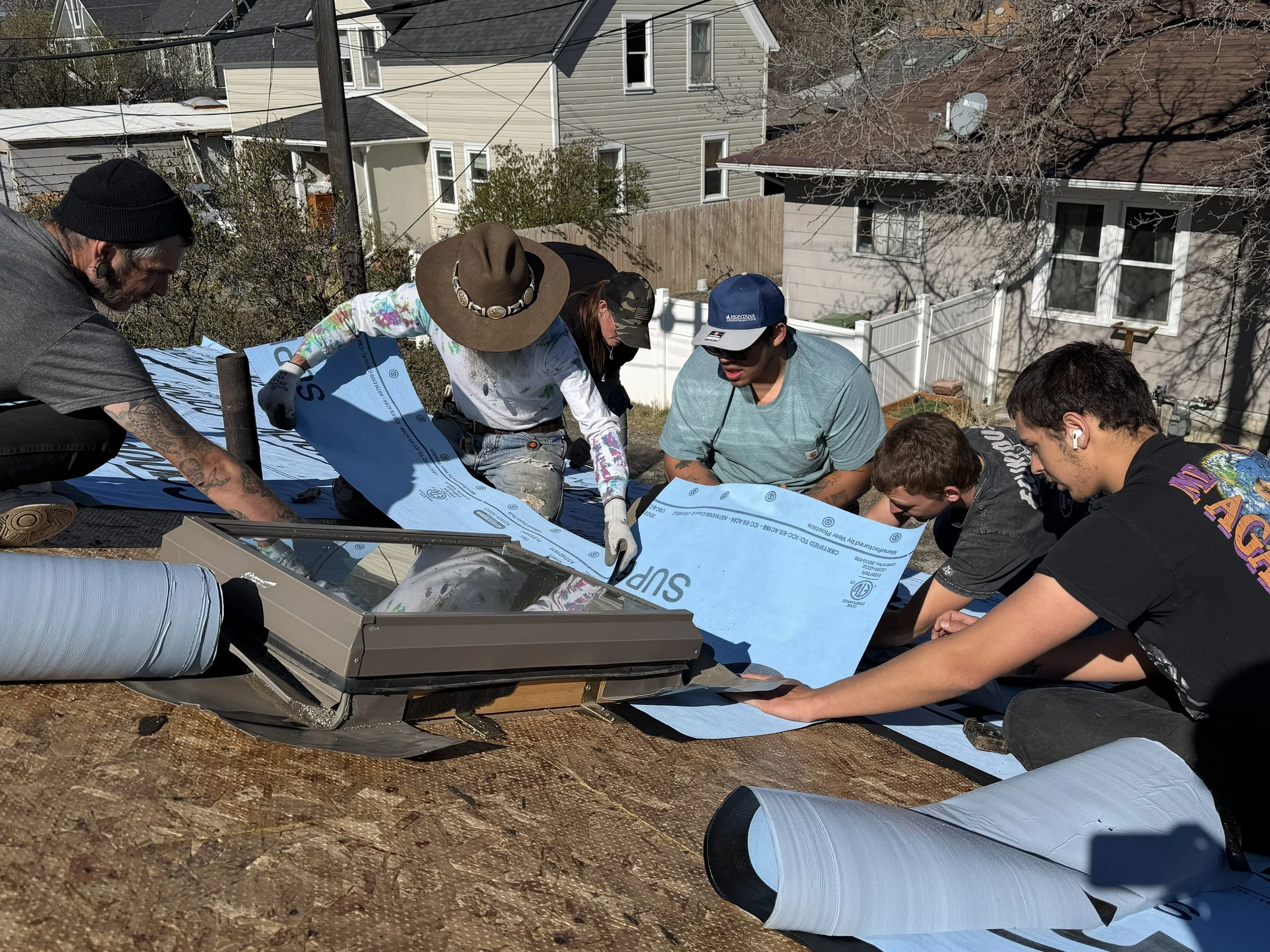 Several people working together on a construction project on a residential roof, handling insulation material surrounded by house and backyard.
