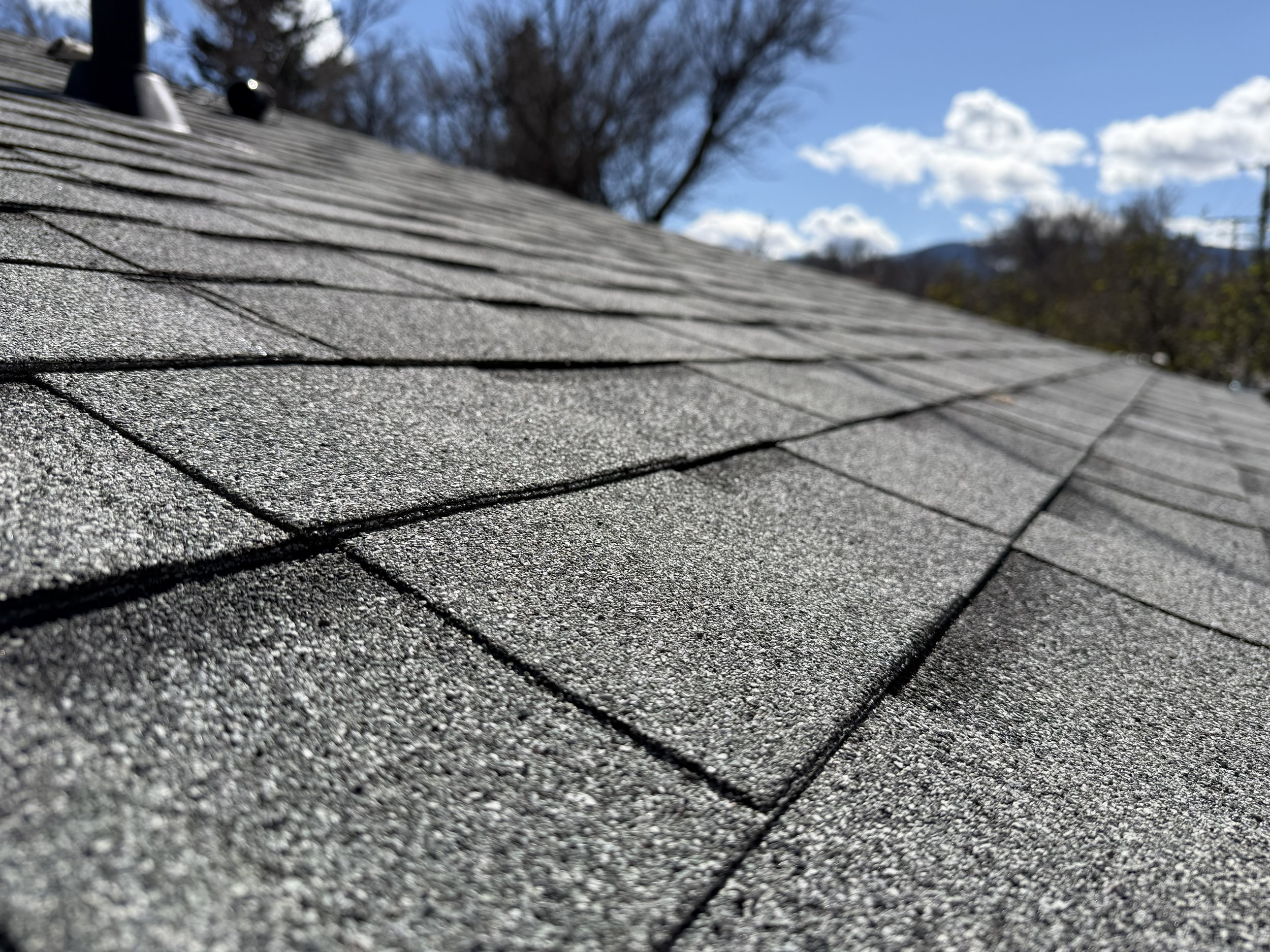 Close-up view of a shingled roof with gray asphalt shingles, with trees and a blue sky with clouds in the background.