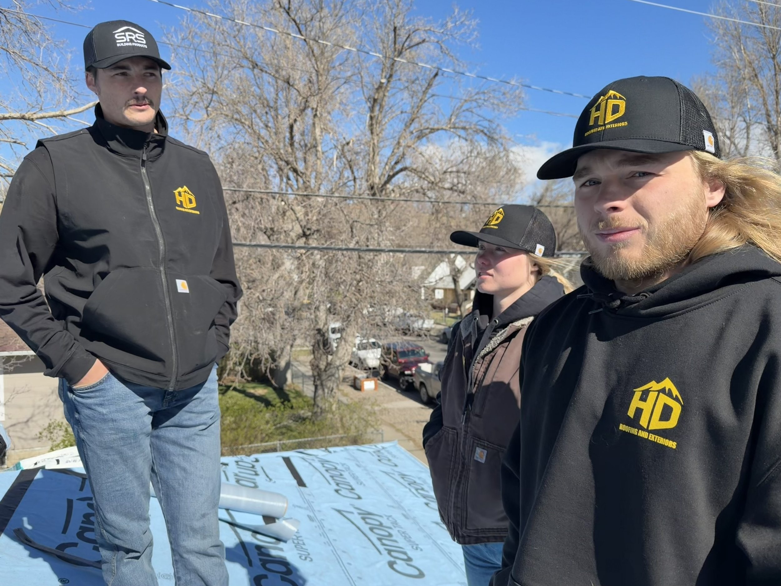 Three men are standing outdoors on a sunny day, wearing black jackets and black baseball caps with logos. They are among roofing materials and banners, with leafless trees and houses in the background.