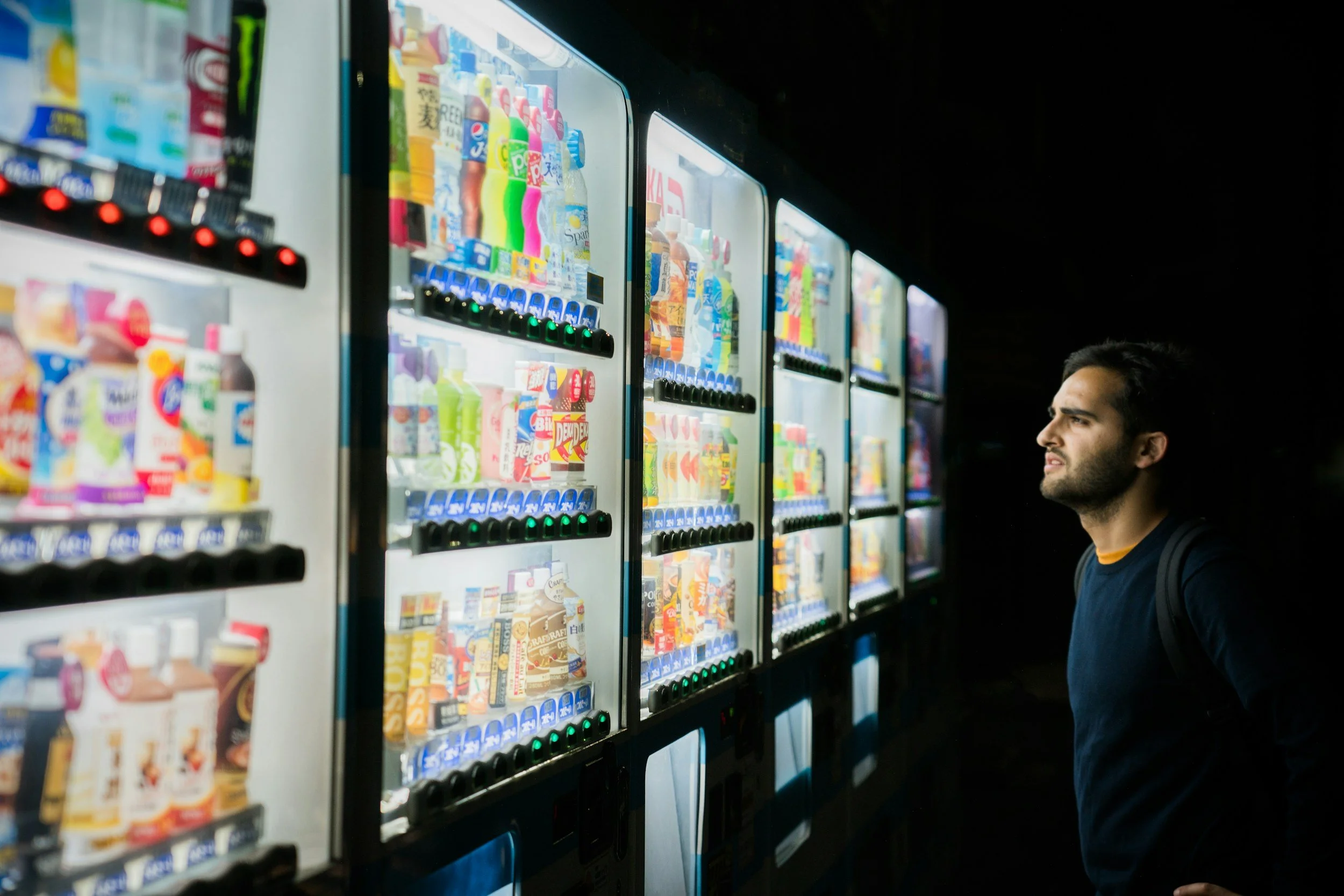 A man with dark hair and a beard, wearing a dark shirt and backpack, looks at vending machines filled with snacks and beverages in a dark environment.