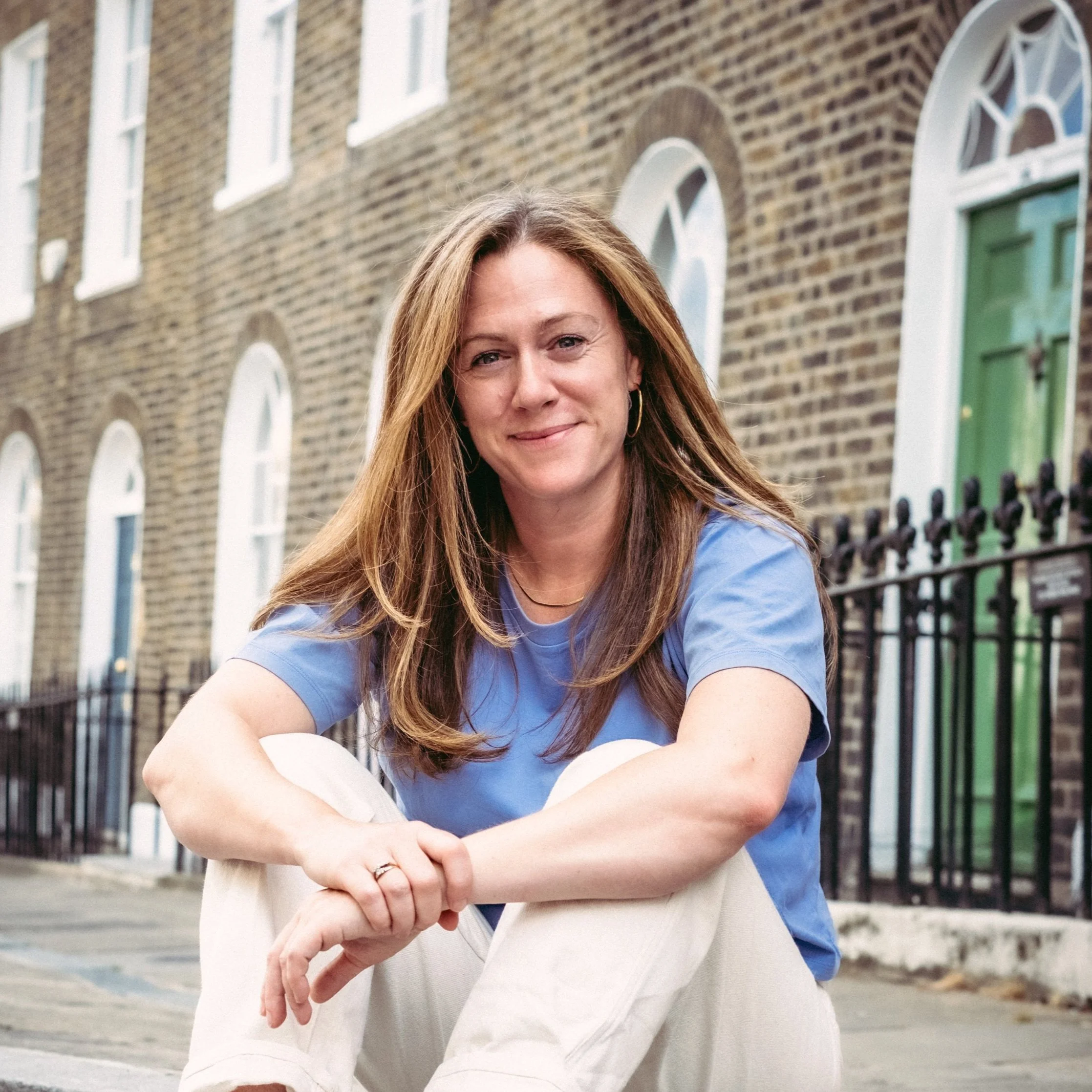 Julia Slay, a woman with long, light brown hair smiles at the famera. She is wearing a light blue t-shirt and white trousers, and sitting down with her hands clasped together. Behind her is a quiet city street. The photo is cropped to a circle.