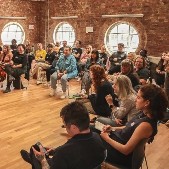 A group of people, of various ages and genders, sit on chairs in a semi-circle. They are looking at a speaker who is out of frame. The photo is cropped to a circle shape.