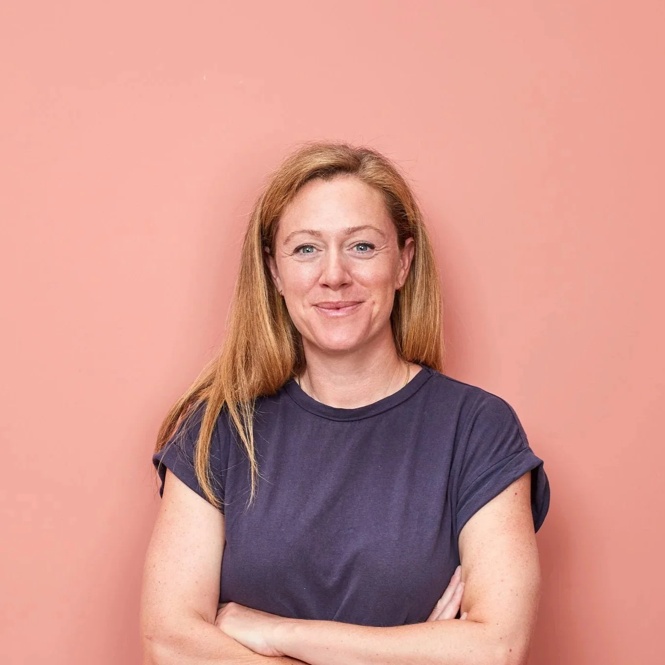 Julia Slay, a woman with long, light brown hair smiles at the famera. She is wearing a dark blue-grey t-shirt and has her arms folded. The photo is a circle shape..