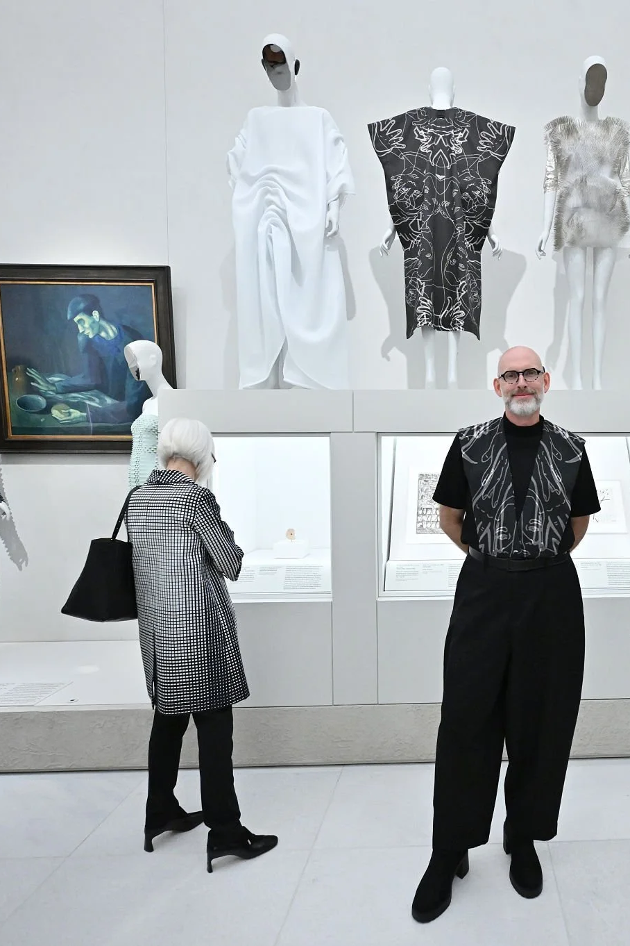 Museum gallery with mannequins dressed in artistic clothing, surrounded by artwork and display cases, and two visitors observing the exhibits.