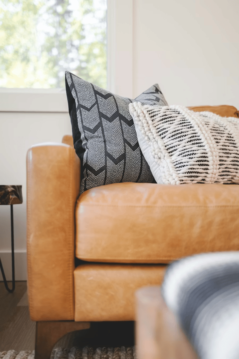 Close-up of a tan leather sofa with decorative cushion pillows and a side table in a bright living room with a window and greenery outside.