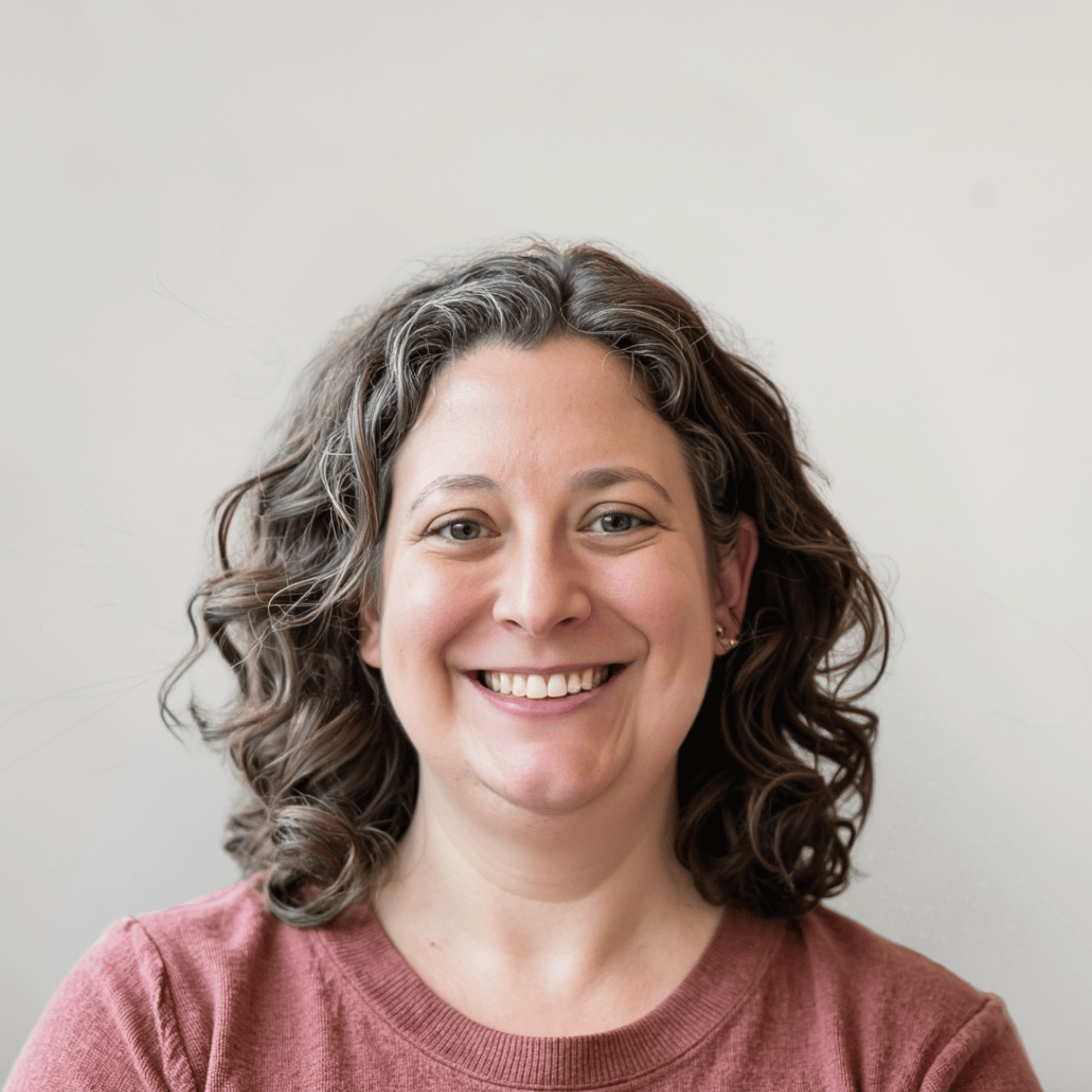 A woman with curly brown hair and a wide smile, wearing a maroon top, standing against a plain light-colored background.