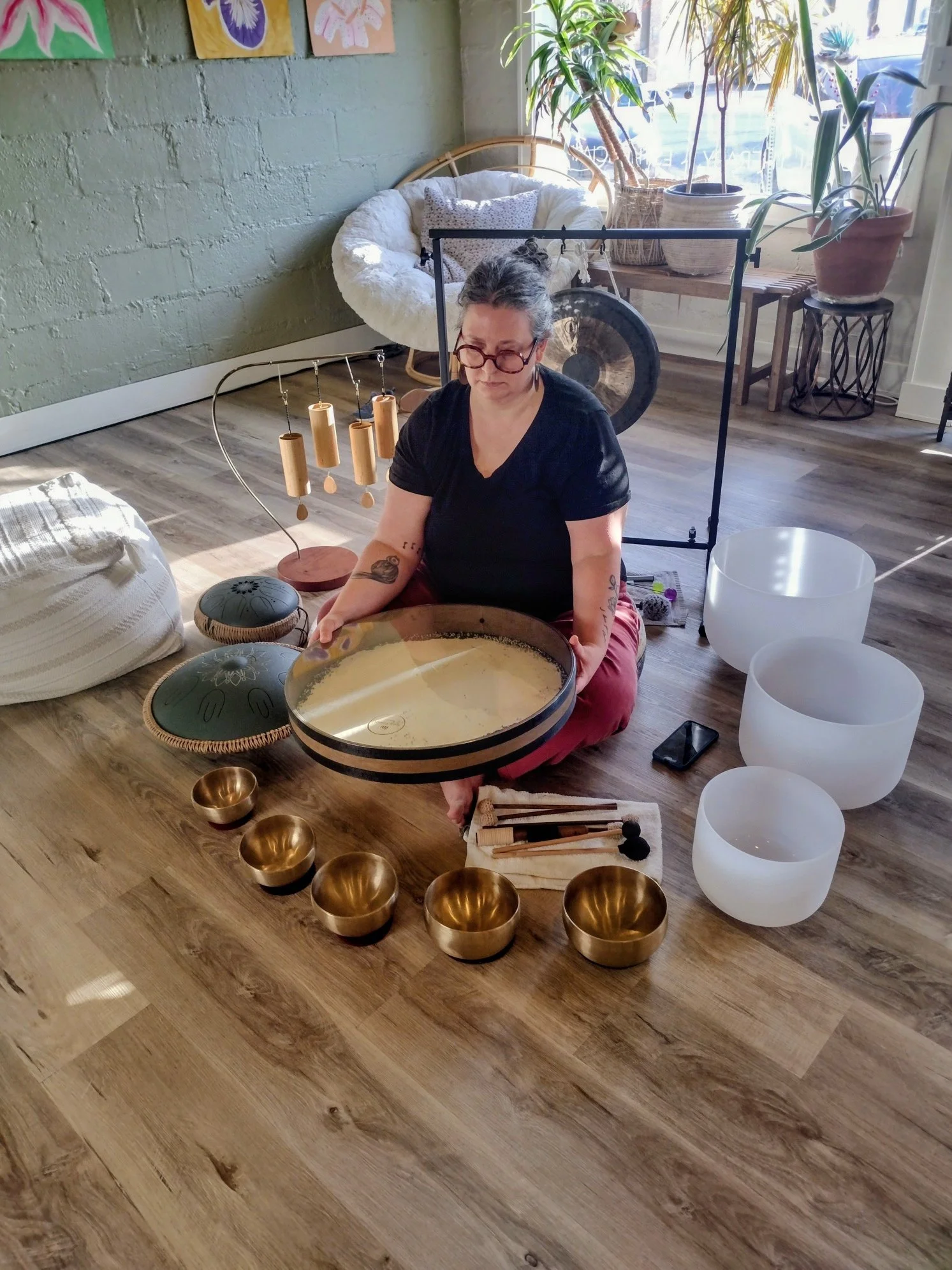 Joann Schermerhorn, founder of Carried Sound, seated on the floor surrounded by crystal singing bowls, brass bowls, a gong, and sound healing instruments at her Overland Park, Kansas City studio