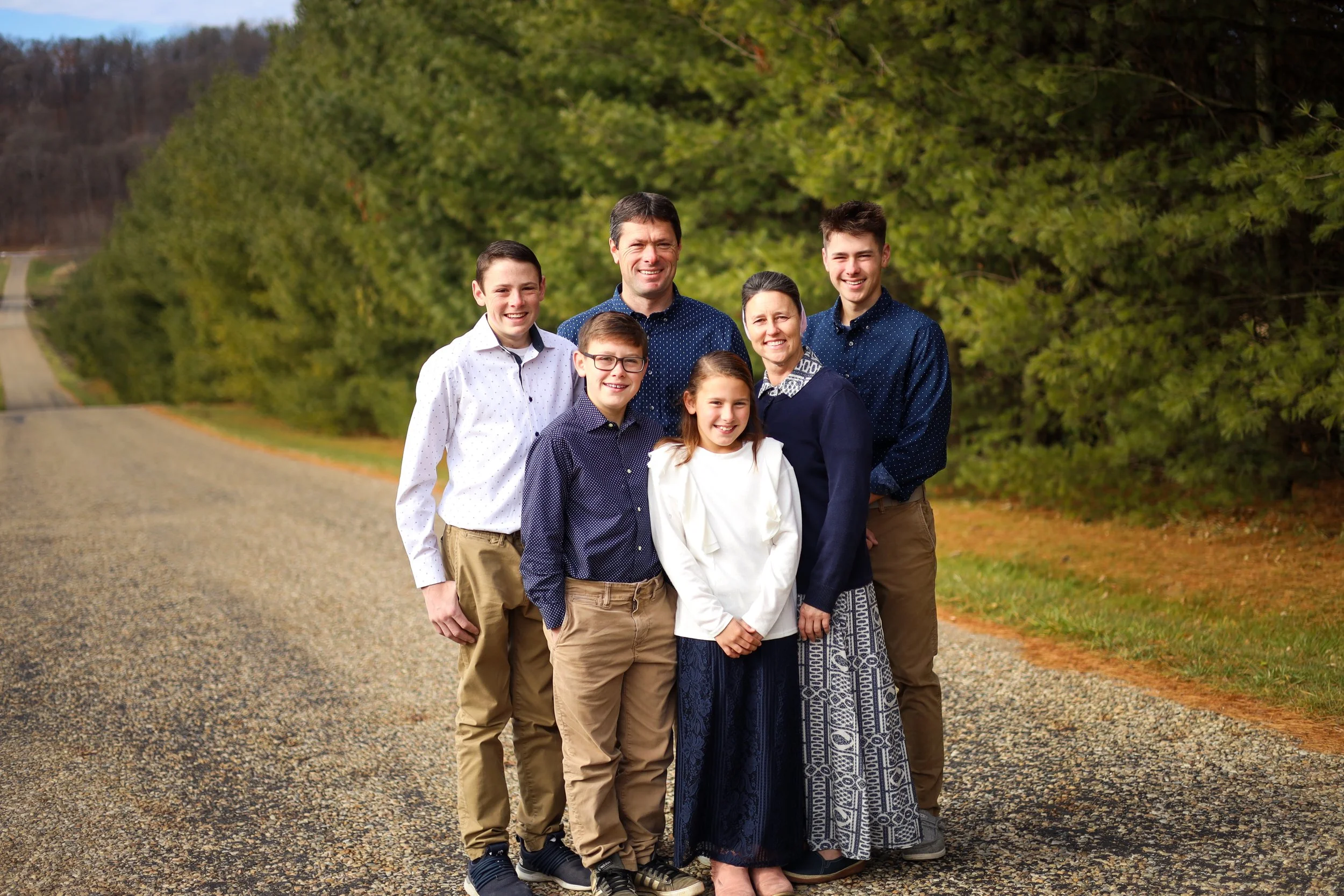 A family of seven standing outdoors on a gravel road with green trees in the background.