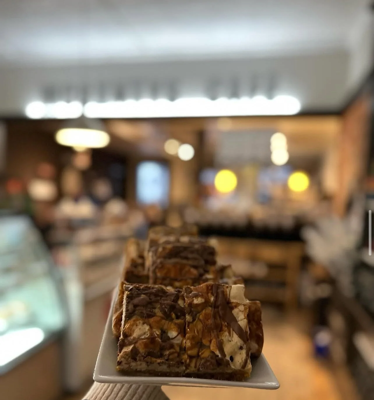 A hand holding a small square plate with caramel and chocolate squares in a bakery or cafe setting, with blurred background of display cases and warm lighting.