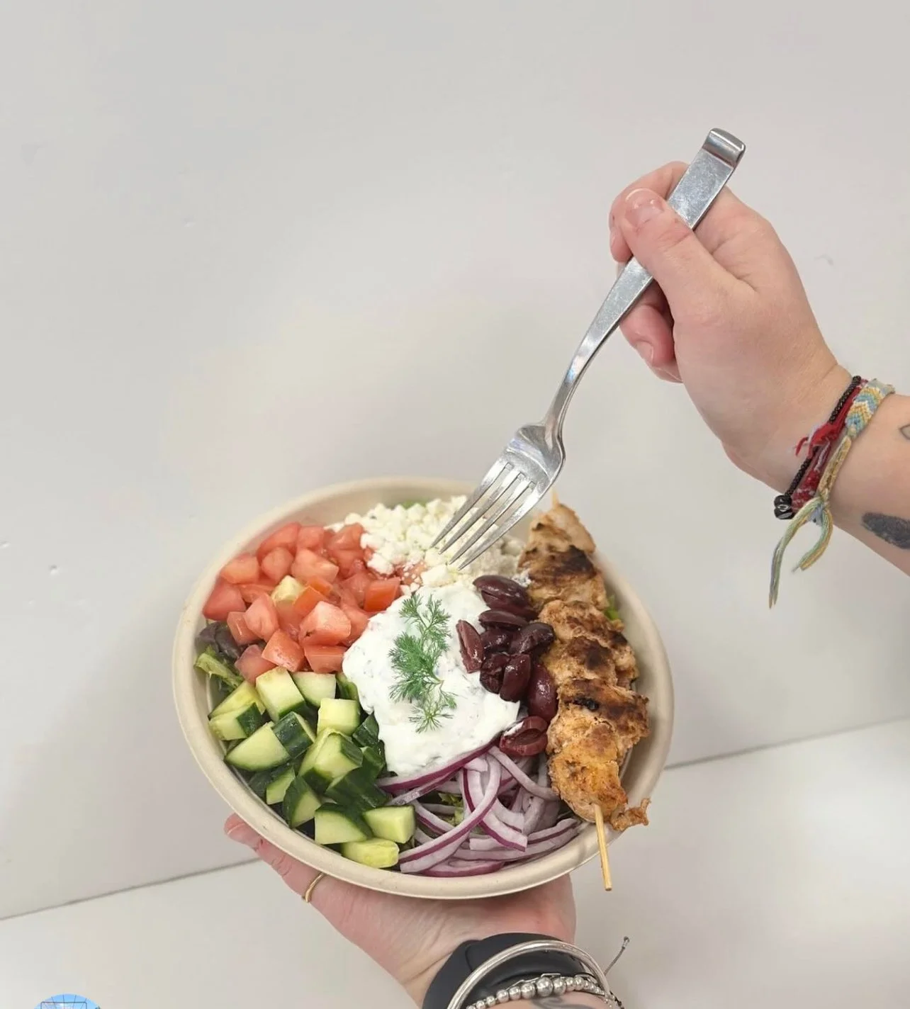 A person holding a bowl of Greek salad with toppings like cucumbers, tomatoes, red onion, black olives, feta cheese, and a grilled chicken skewer, garnished with fresh herbs.