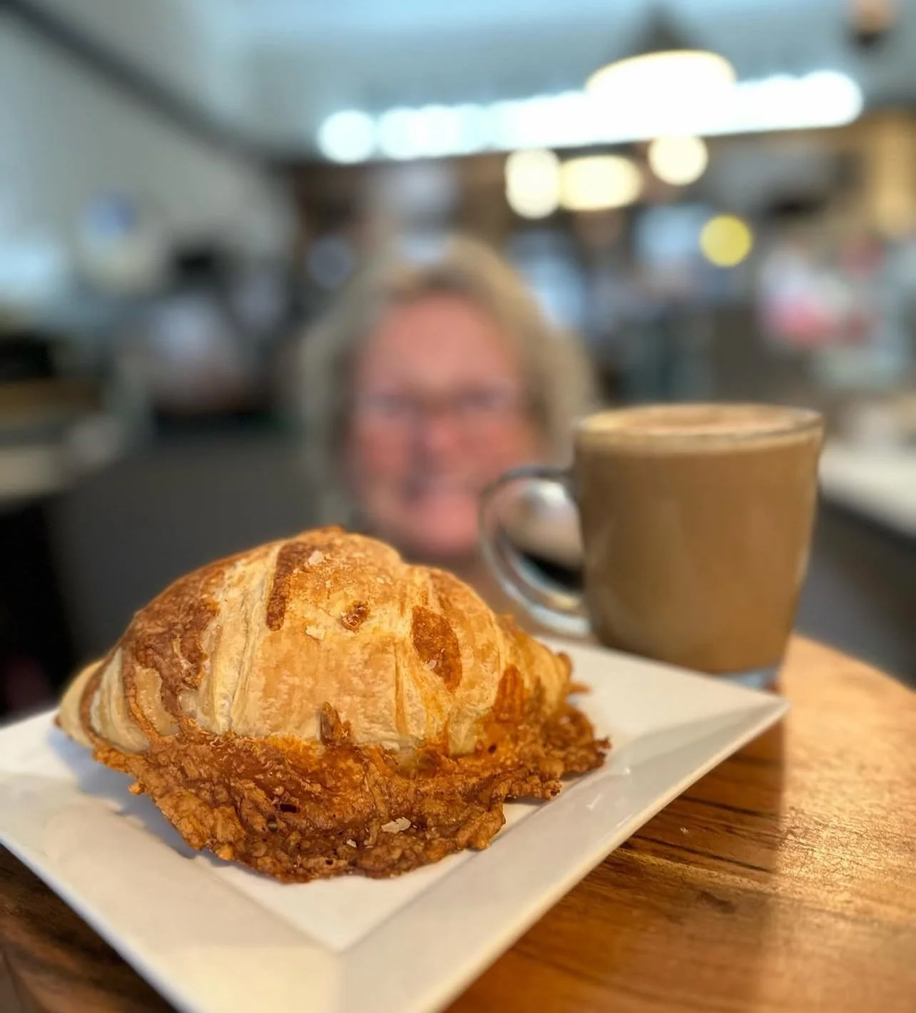 A plate with a croissant and a mug of coffee on a wooden table in a cafe, with a smiling woman in the background.