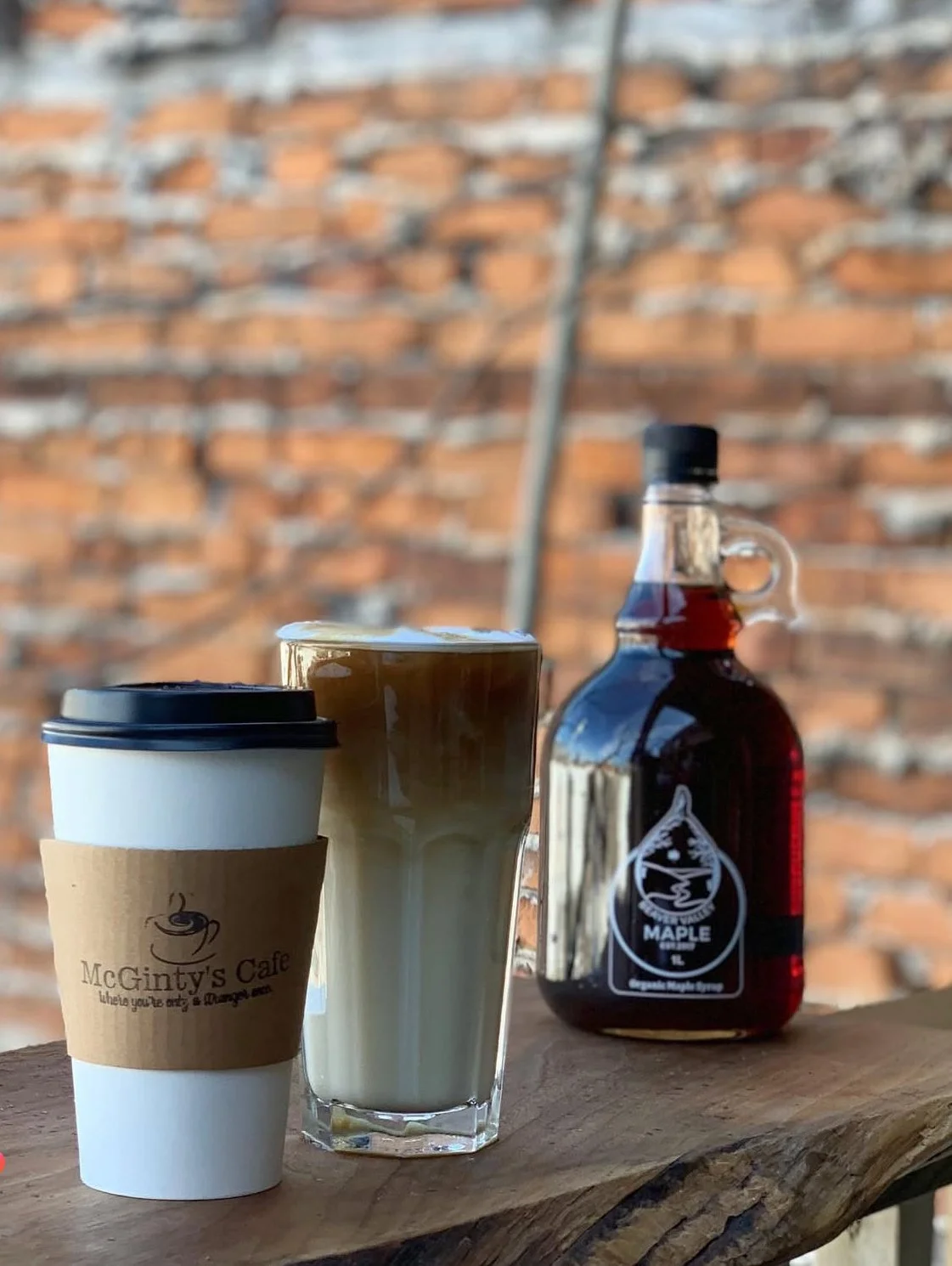 A to-go coffee cup, a glass of coffee with foam, and a syrup bottle labeled 'MAPLE' sit on a wooden surface against a brick wall background.