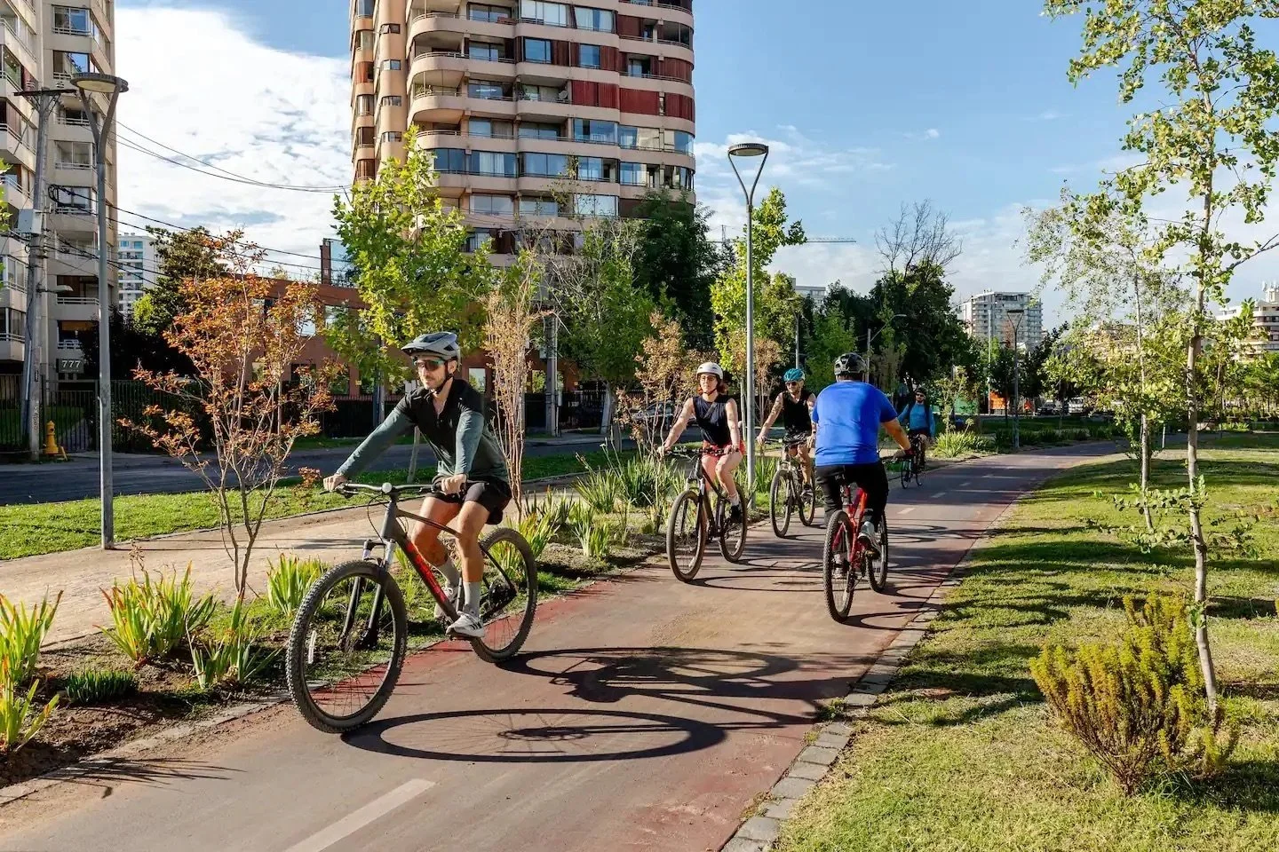 People riding bicycles on a paved path in an urban park with trees, grass, and apartment buildings in the background under a partly cloudy sky.
