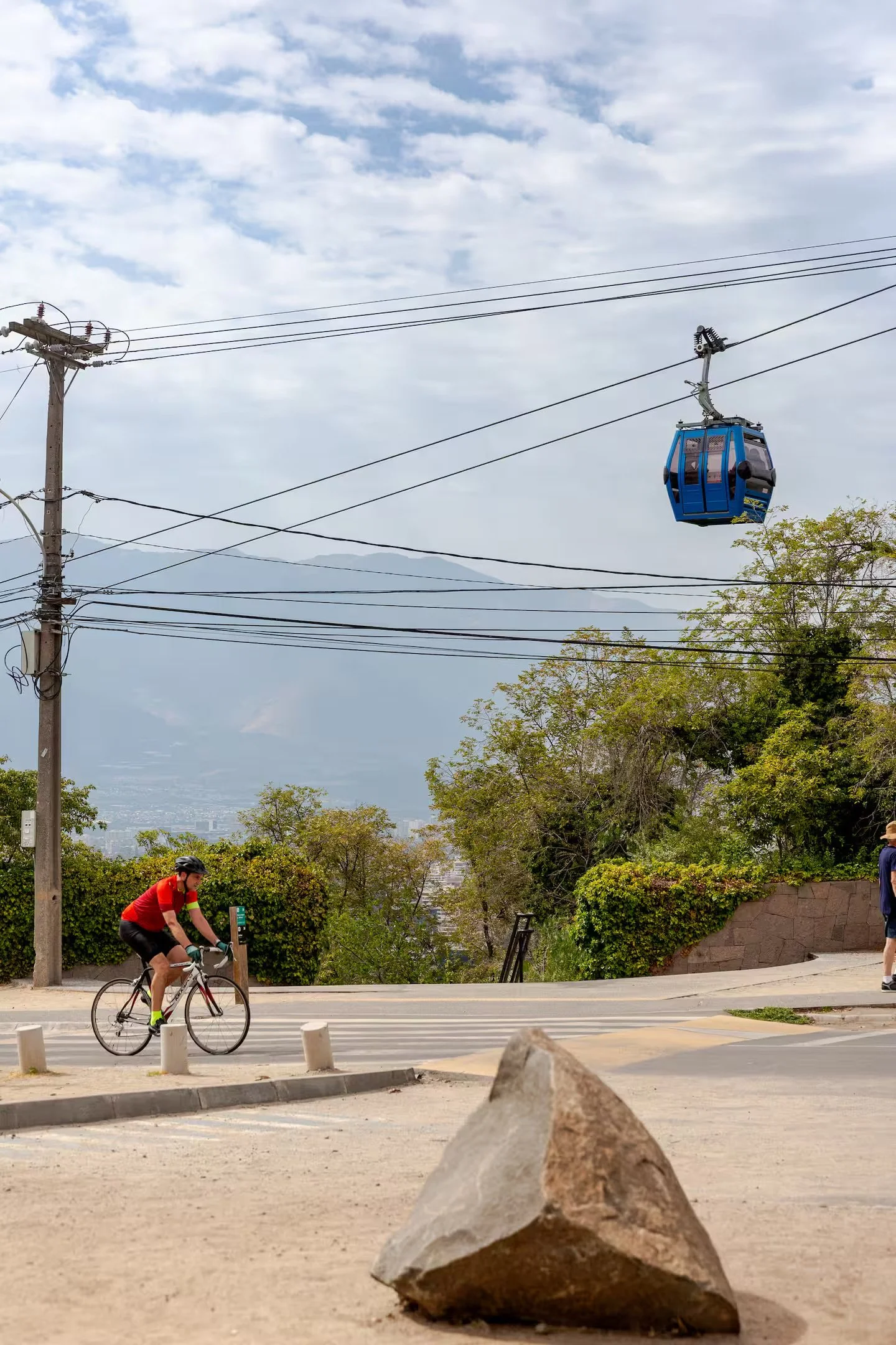 A person in a red shirt riding a bicycle on a street with trees and a mountain in the background. A large rock in the foreground and a blue cable car suspended in the sky.