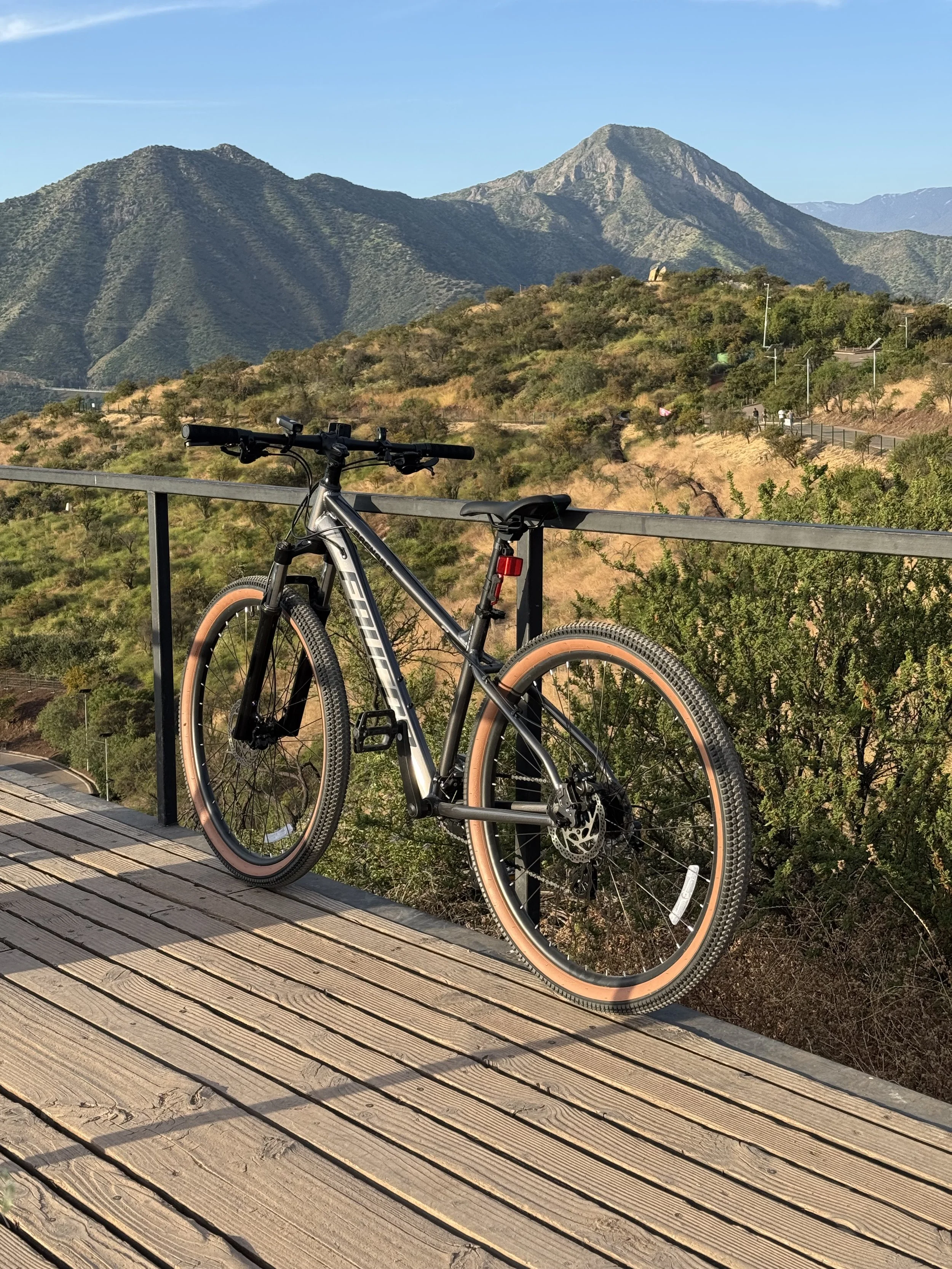 A black mountain bike with tan tires leaning against a railing on a wooden deck, overlooking a hilly landscape with mountains in the background on a clear sunny day.