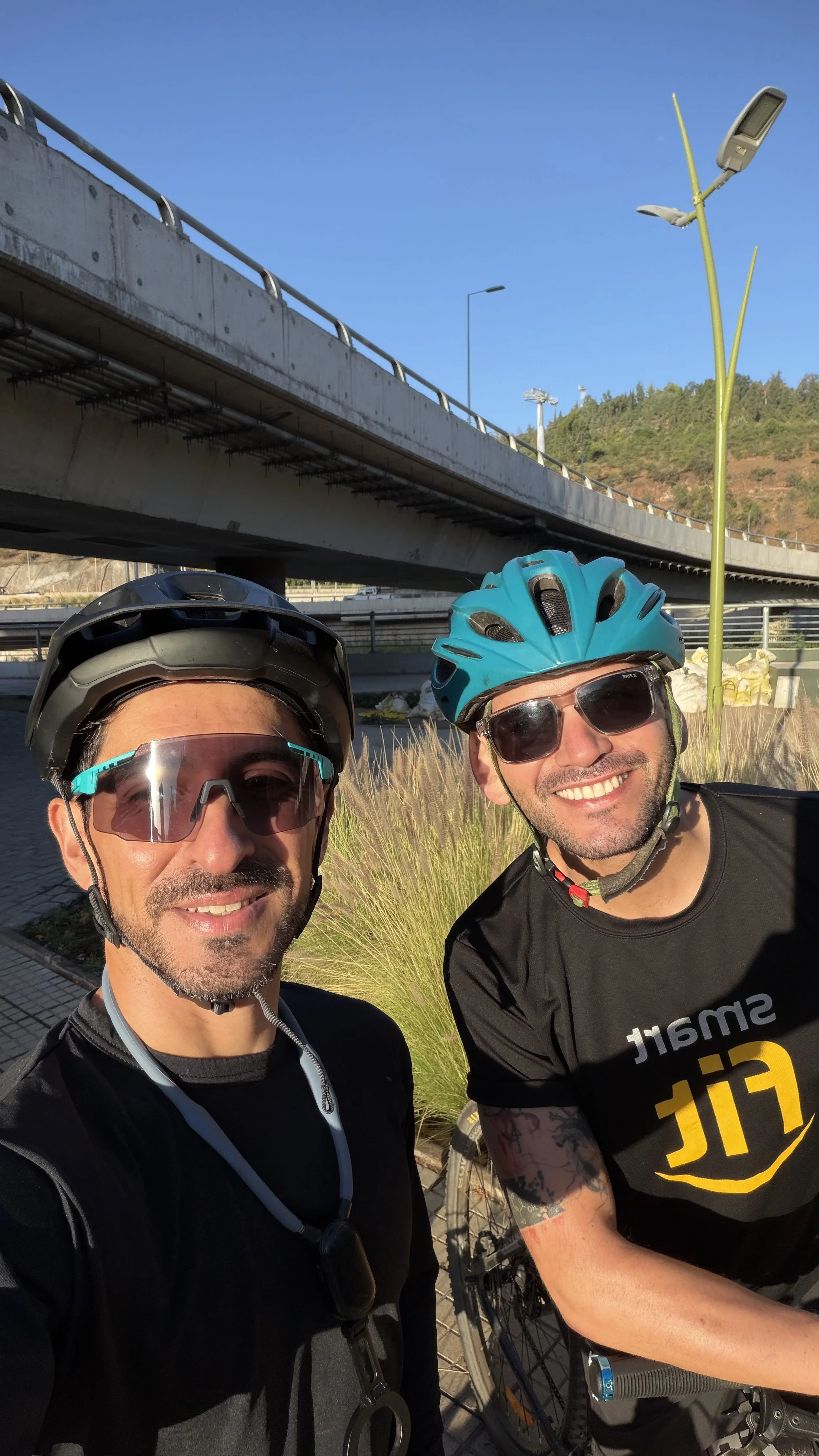 Two men wearing cycling helmets and sunglasses taking a selfie outdoors under a bridge, with greenery and a hill in the background.