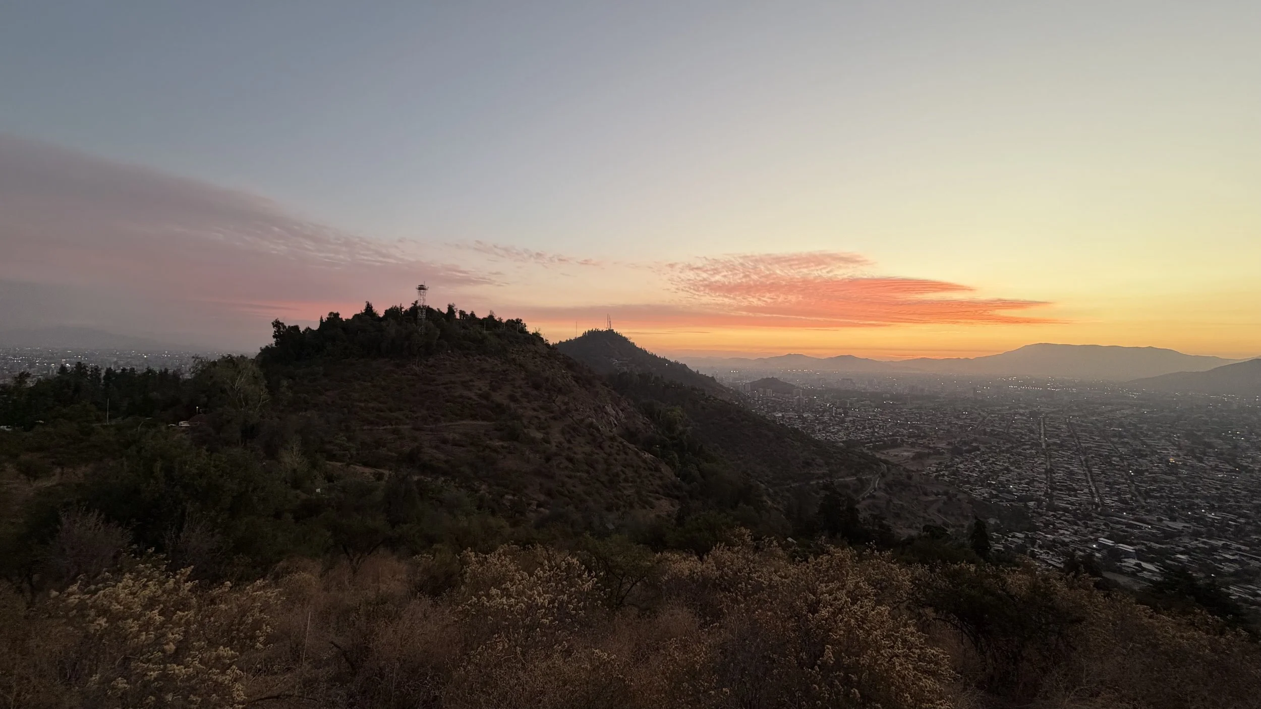 Sunset over a hilly cityscape with a mountain in the background, showing a colorful sky with pink, orange, and yellow hues.