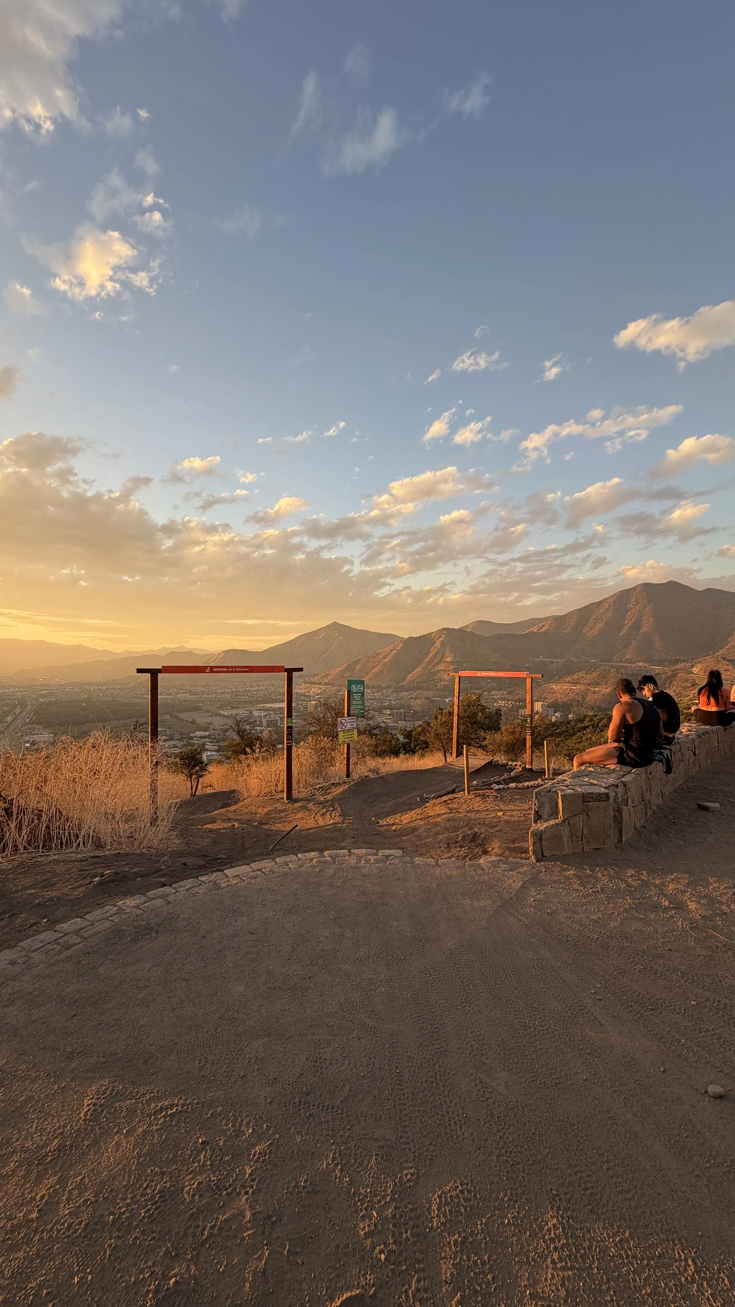 People sitting on a stone wall during sunset at a mountain overlook, with two empty sign frames in the foreground and mountains in the distance.