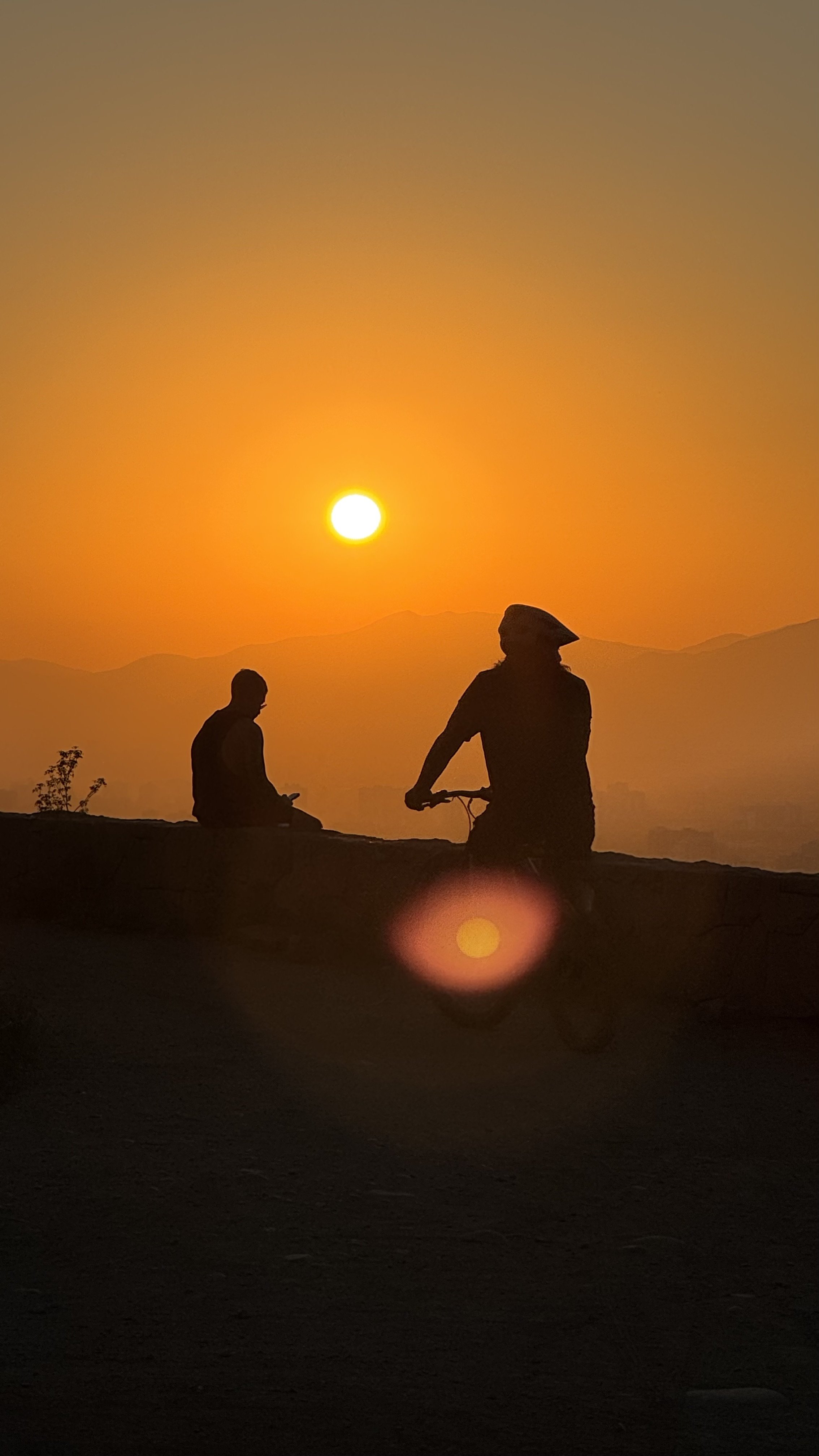 Silhouettes of two people, one on a bicycle and another sitting on a ledge, against a vibrant orange sunset with mountains in the background.