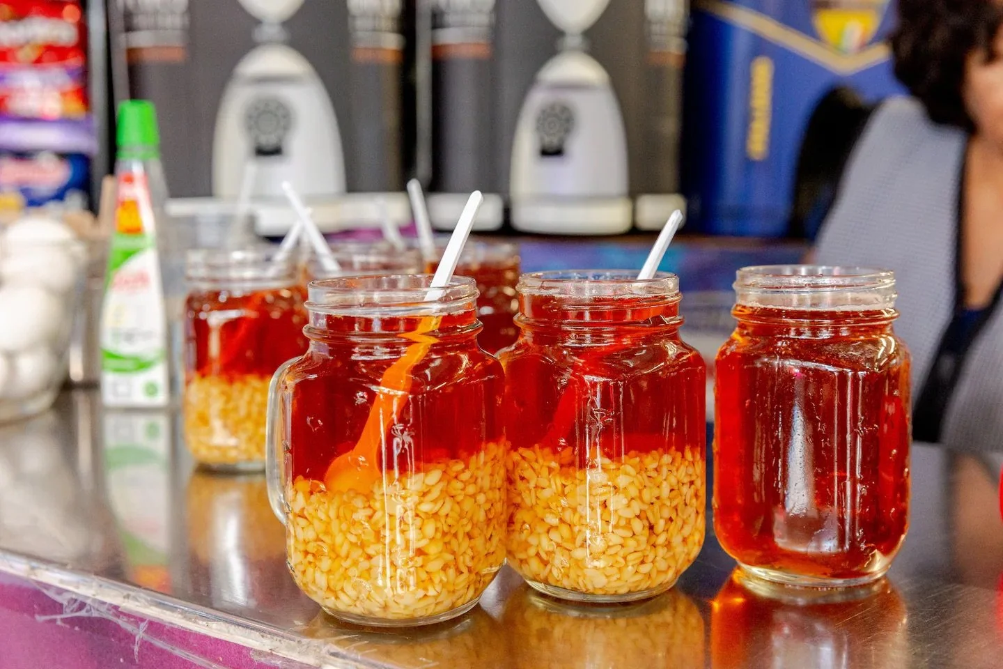 Four mason jars filled with red liquid, containing yellow chili peppers at the bottom, placed on a metal counter with stir sticks inside. In the blurred background, there are dispensers and a person partially visible on the right.