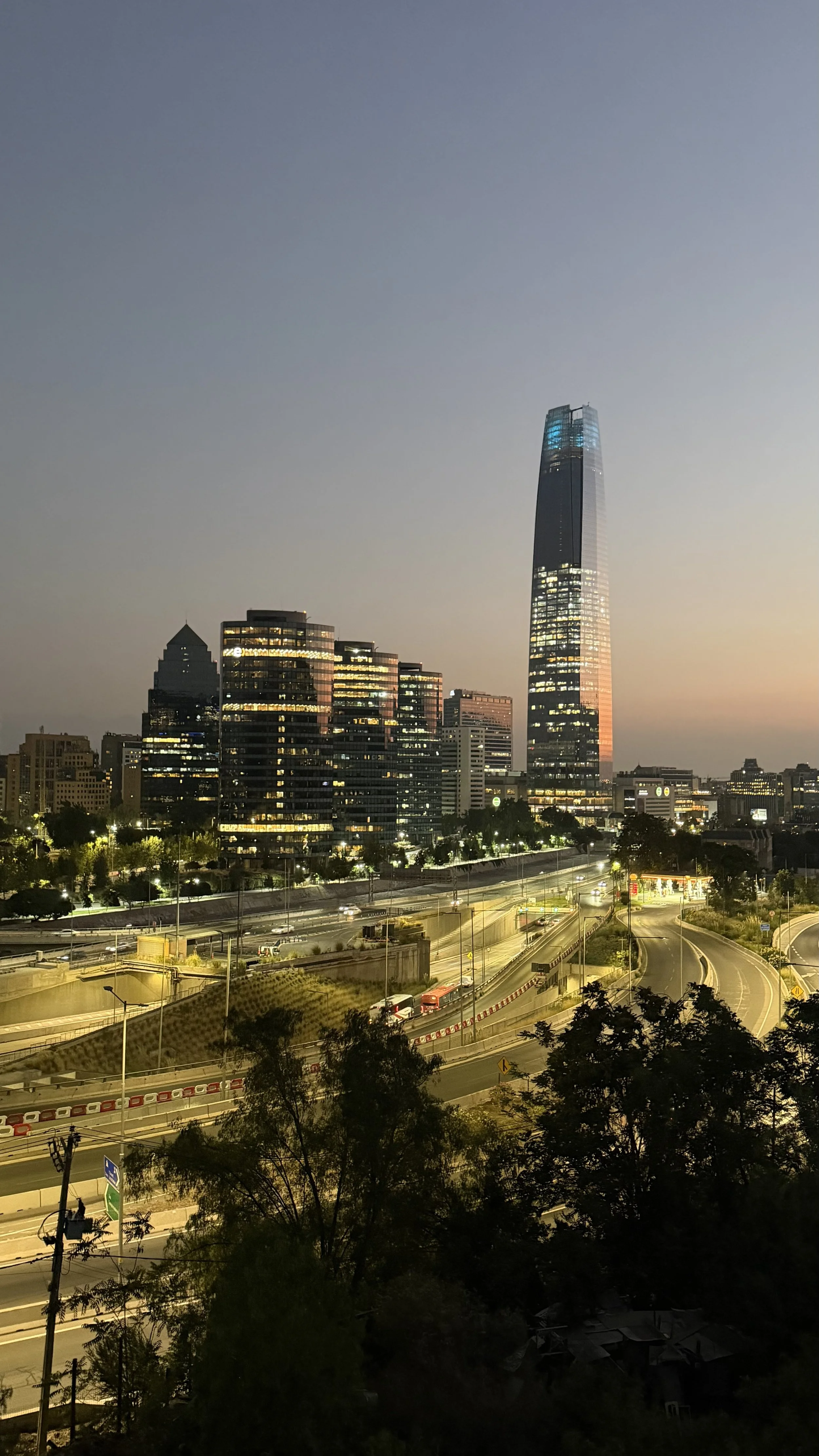 City skyline at dusk featuring tall buildings and a prominent skyscraper, with busy highways and trees in the foreground.