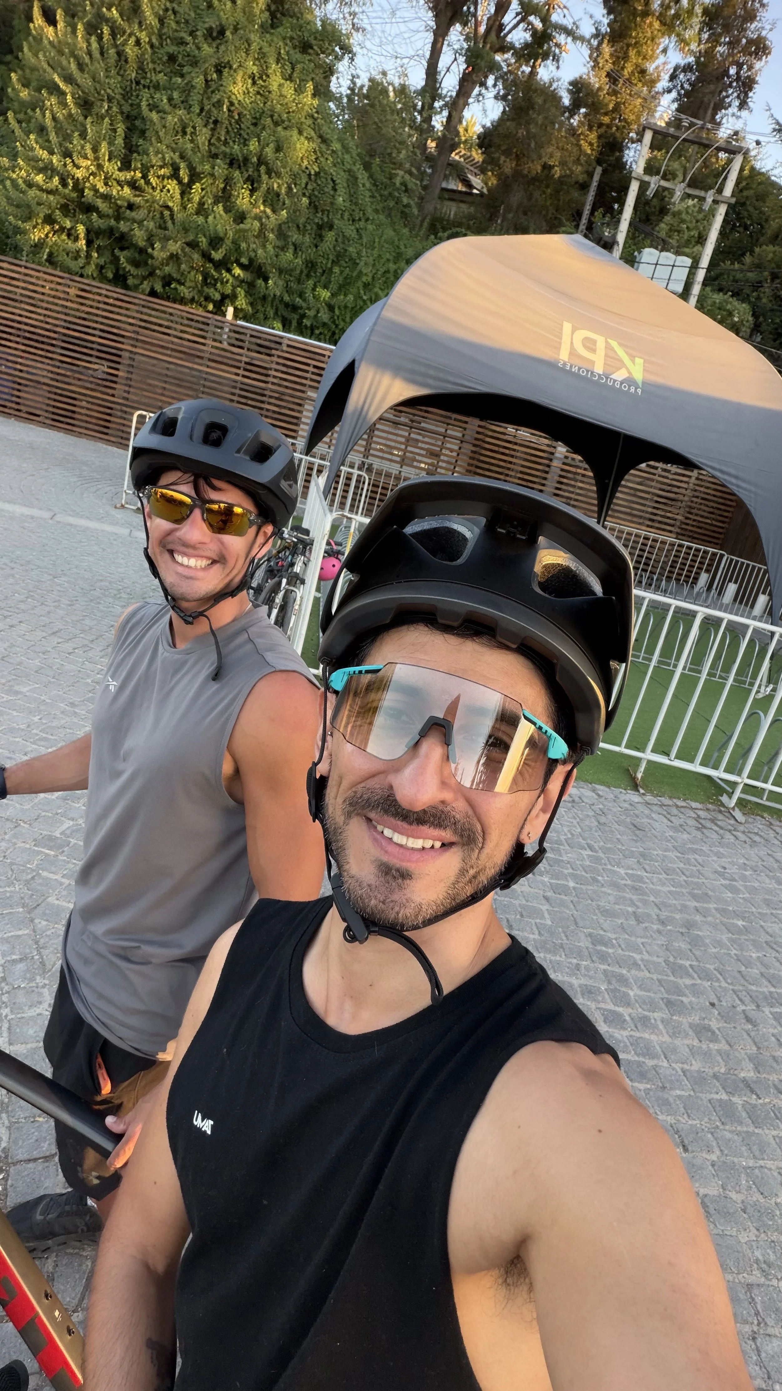 Two men wearing cycling helmets and sunglasses taking a selfie outdoors on a cobblestone path, smiling. One is wearing a sleeveless gray shirt, the other a sleeveless black shirt, with trees, a tent, and a fence in the background.