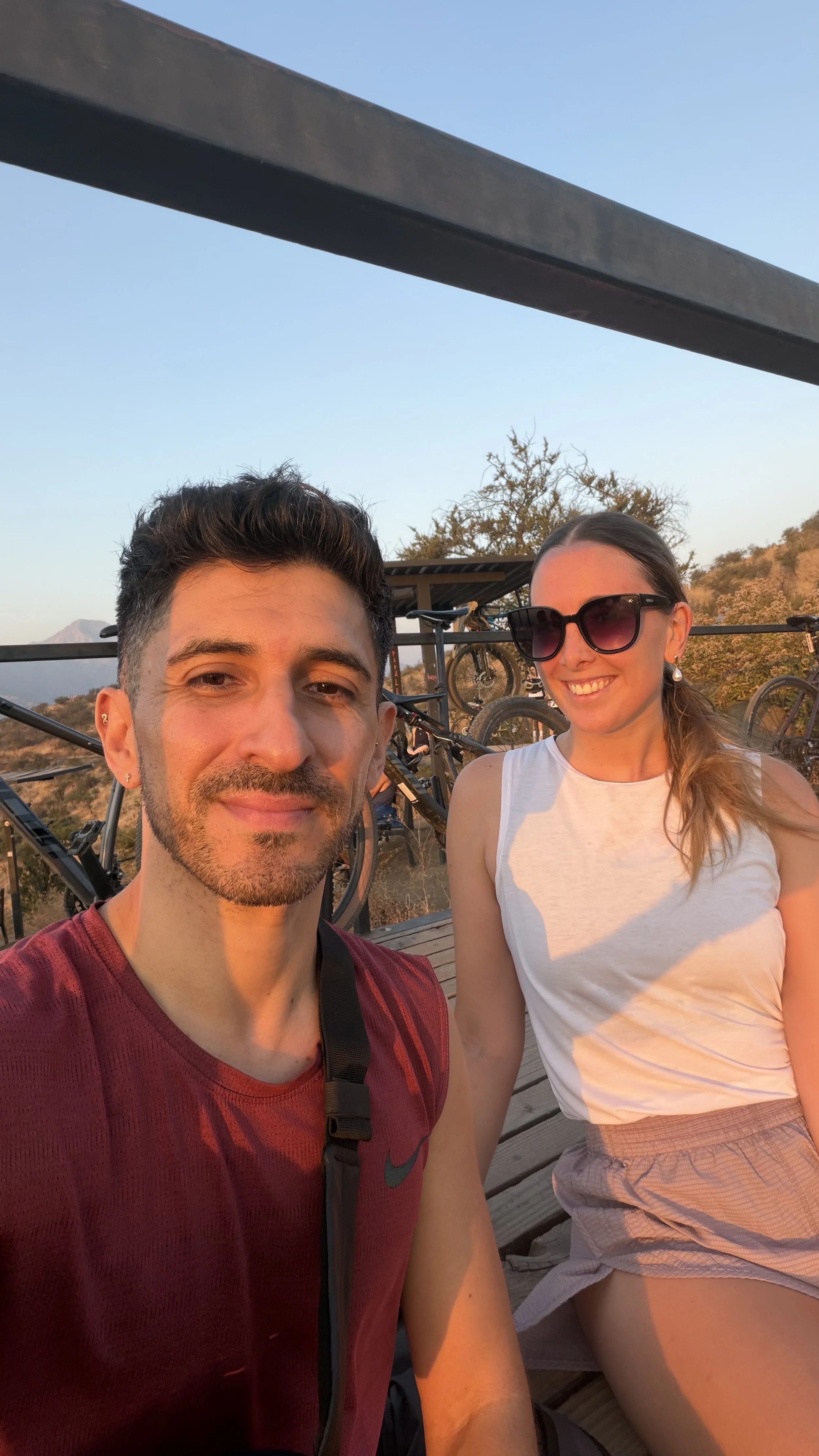 Man and woman taking a selfie outdoors at sunset, with bicycles in the background.