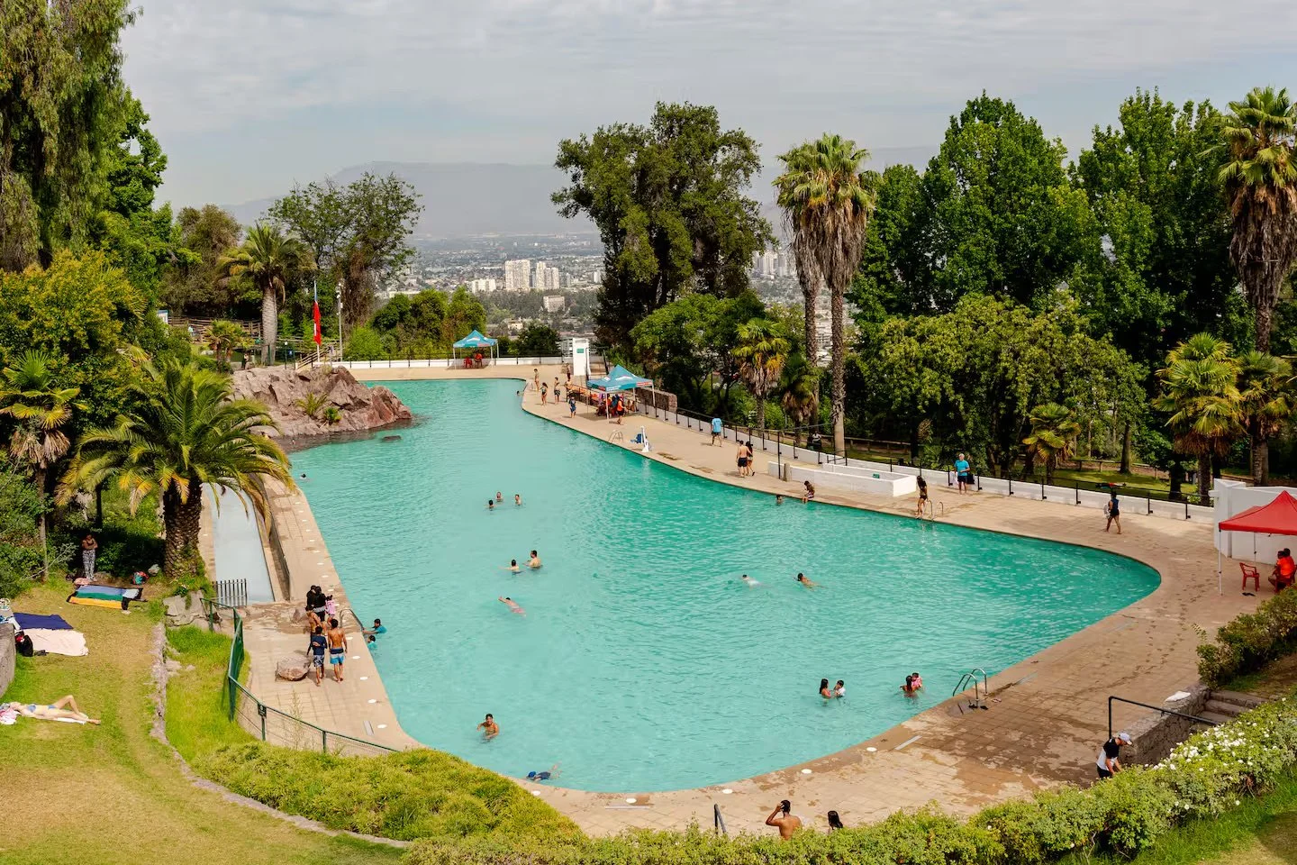 Public outdoor swimming pool surrounded by green trees and people swimming and relaxing.