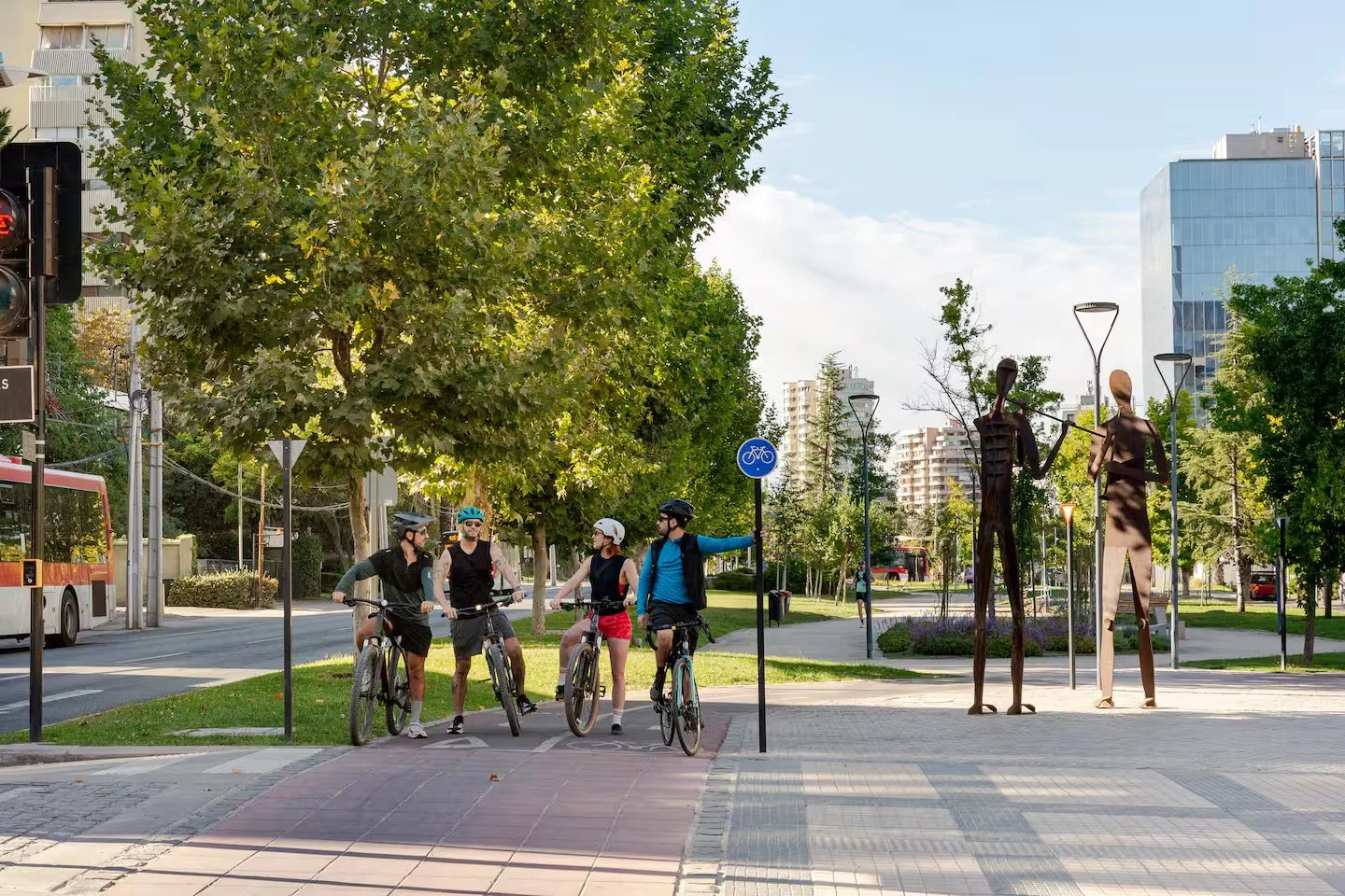 Four cyclists wearing helmets and casual sportswear, standing with their bikes on a city sidewalk near a bike lane sign, engaged in conversation, with three tall abstract metal sculptures of elongated human figures and a background of green trees, buildings, and a clear sky.