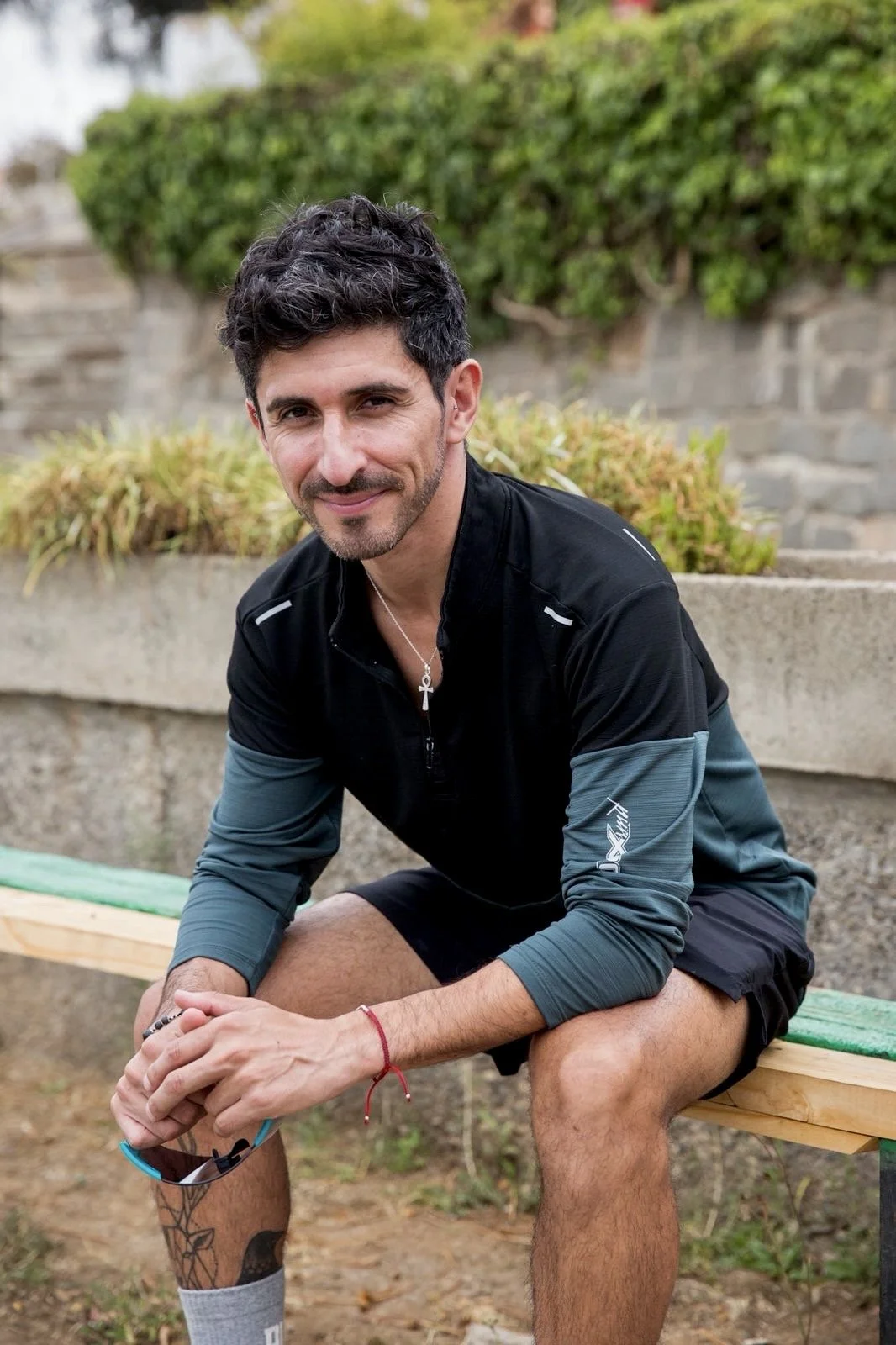 A man with dark curly hair, a beard, and medium skin tone sitting on a green and wooden bench outdoors, smiling at the camera. He is wearing a black and gray long-sleeve athletic shirt, black shorts, gray socks, and has a cross necklace. There is greenery and a stone wall in the background.
