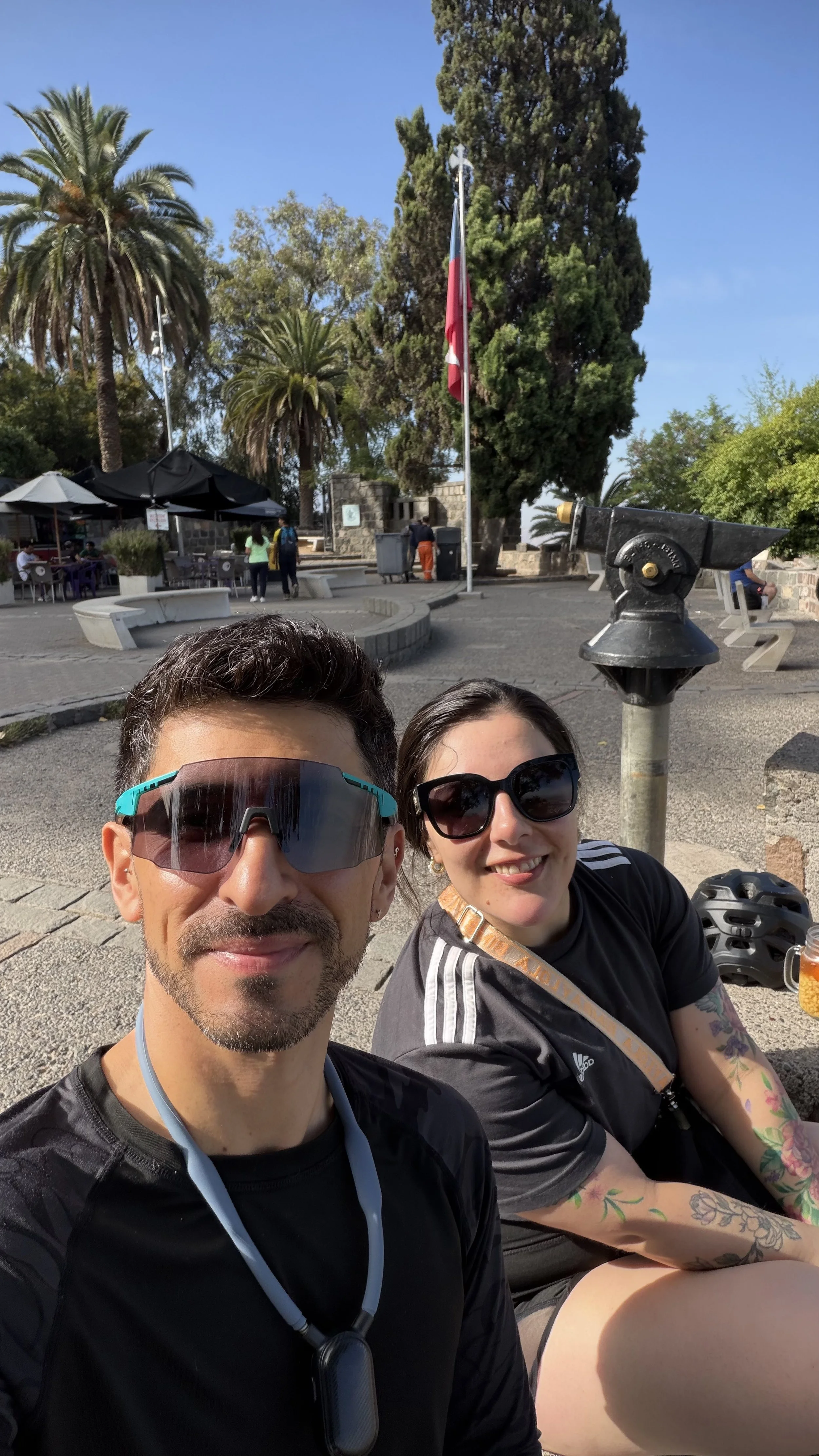A man and woman sitting outdoors in a park, taking a selfie with trees, a flag, and a coin-operated viewing telescope in the background.