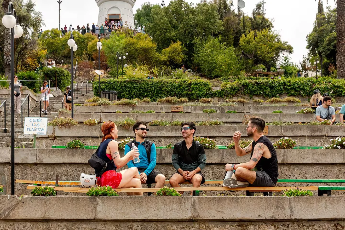 Four people sitting on a bench in an outdoor staircase park, talking and laughing, with many other people walking around in the background and on the upper level. There are trees, bushes, and park lamps.