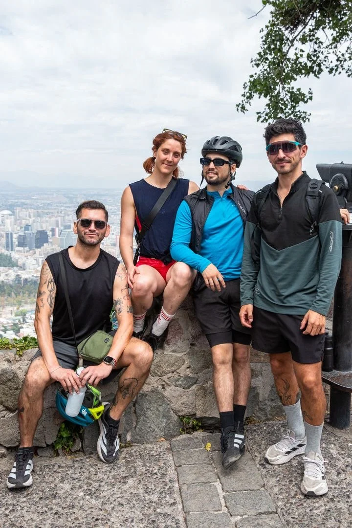 Four friends in athletic clothing and sunglasses, one with a bike helmet, posing on a stone ledge with a cityscape in the background, indicating they are outdoors in an urban setting.