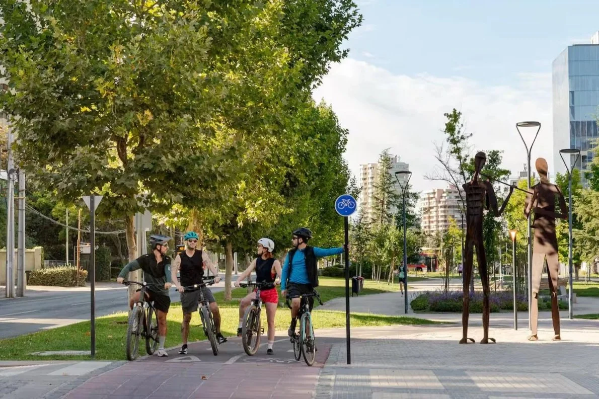 Four people with bicycles wearing helmets, standing on a paved park path, talking to each other. The park has trees, grass, and modern sculptures of two tall, abstract human figures. A blue bicycle lane sign is visible, and city buildings are in the background.