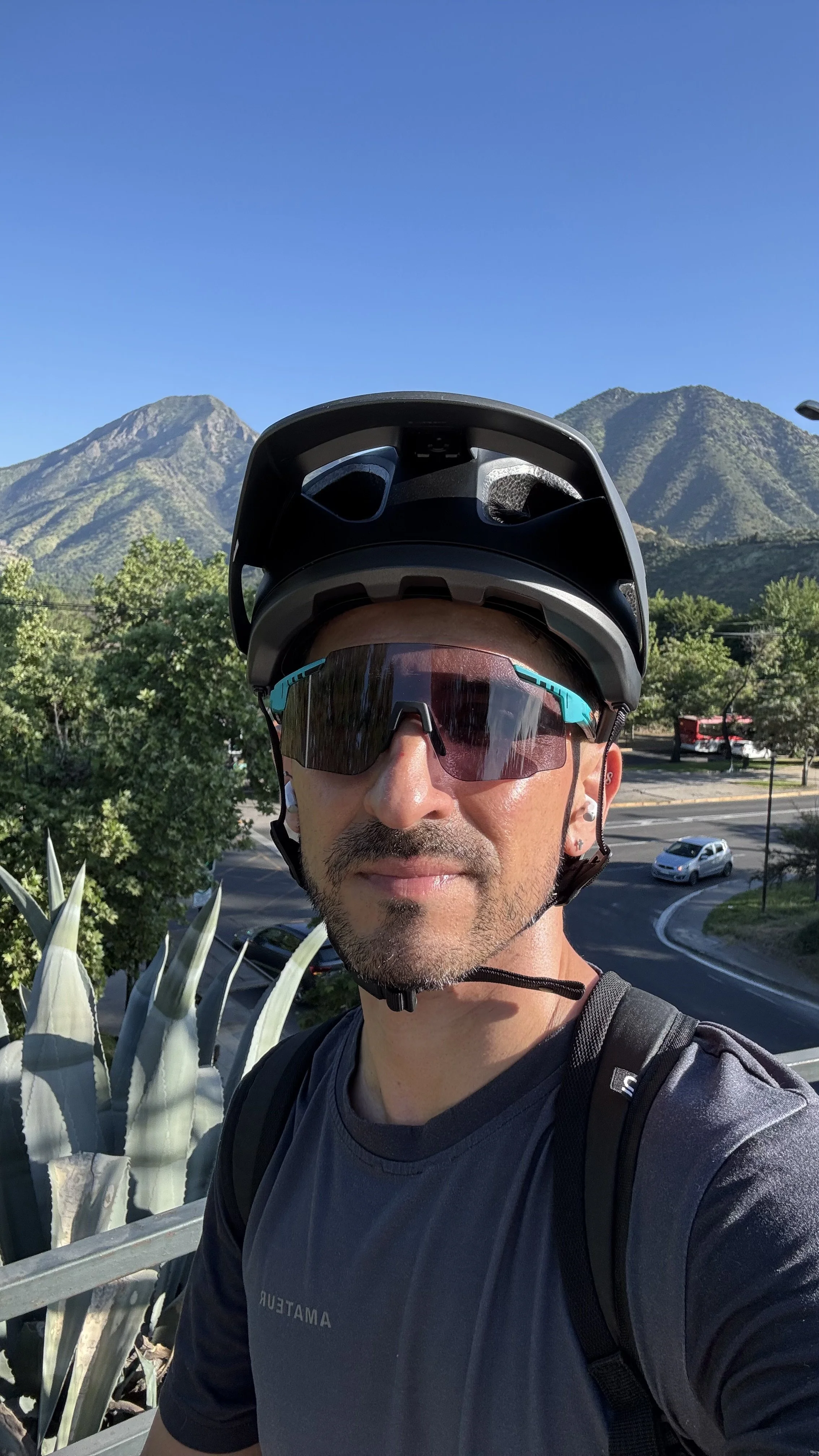 A man wearing a helmet and sunglasses taking a selfie outdoors with mountains and a clear blue sky in the background.