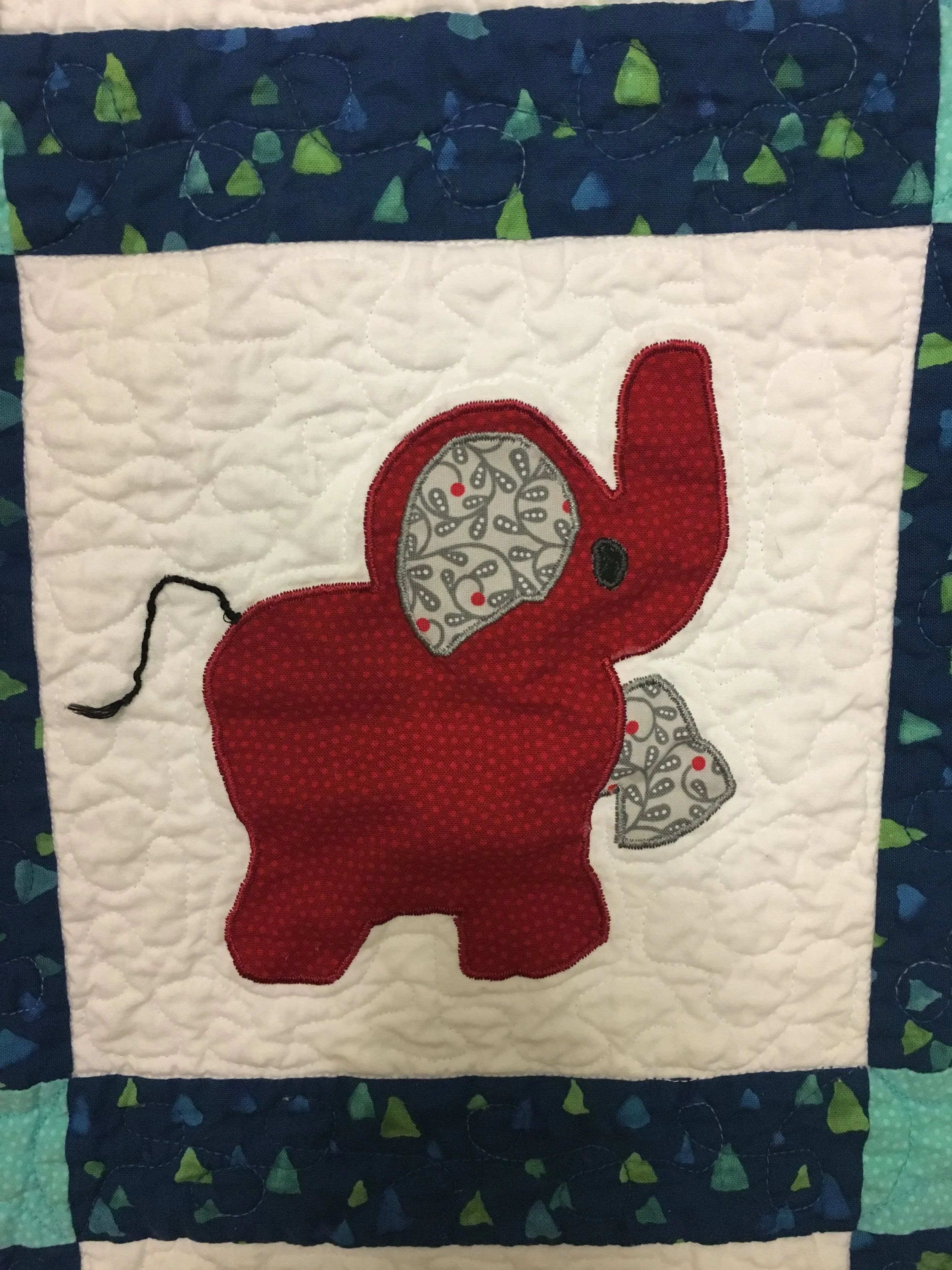 A quilted fabric piece featuring an applique of a red elephant with grey and white patterned ears on a white background, surrounded by a multicolored border.