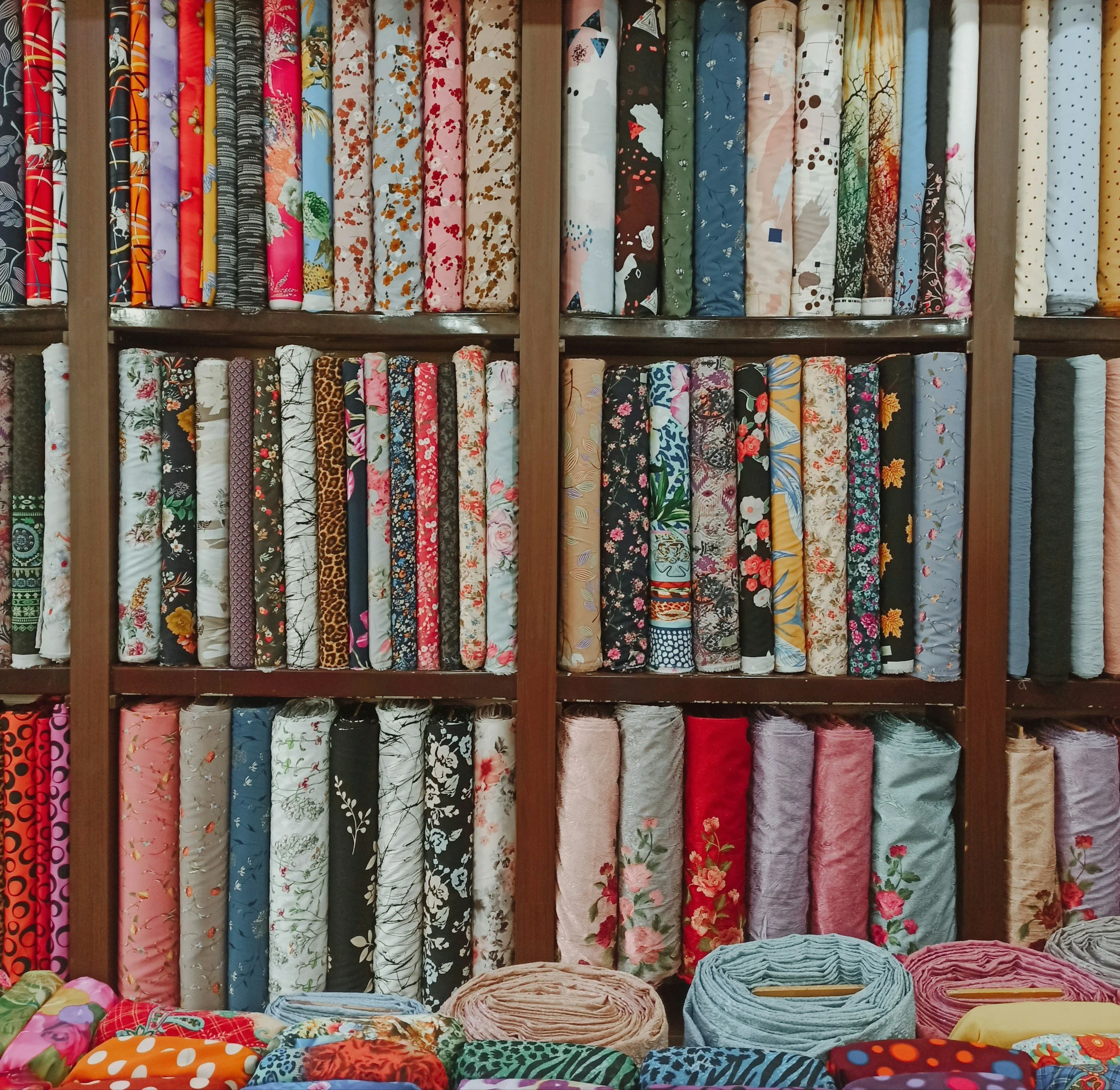 A wall of shelves filled with rolled fabric bolts featuring various colorful patterns, with some fabric bolts laid out on a table at the bottom.