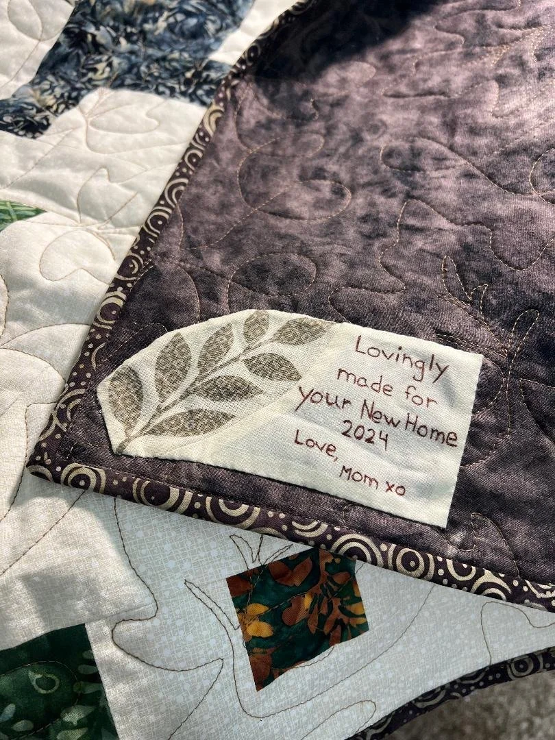 Close-up of a quilt with a handwritten note embroidered on a white fabric square that says, 'Lovingly made for your New Home 2024 Love, Mom xo.' The quilt features various fabric patterns and quilting stitches.