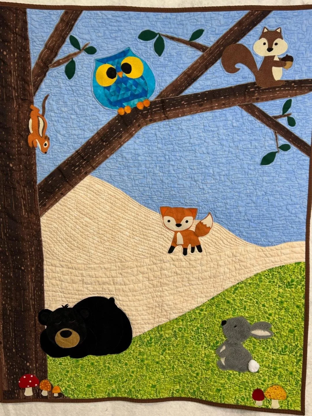 A quilt depicting a woodland scene with animals: a black bear, a gray rabbit, a fox, an owl, a squirrel, and a chipmunk on a tree. There are mushrooms on the ground.