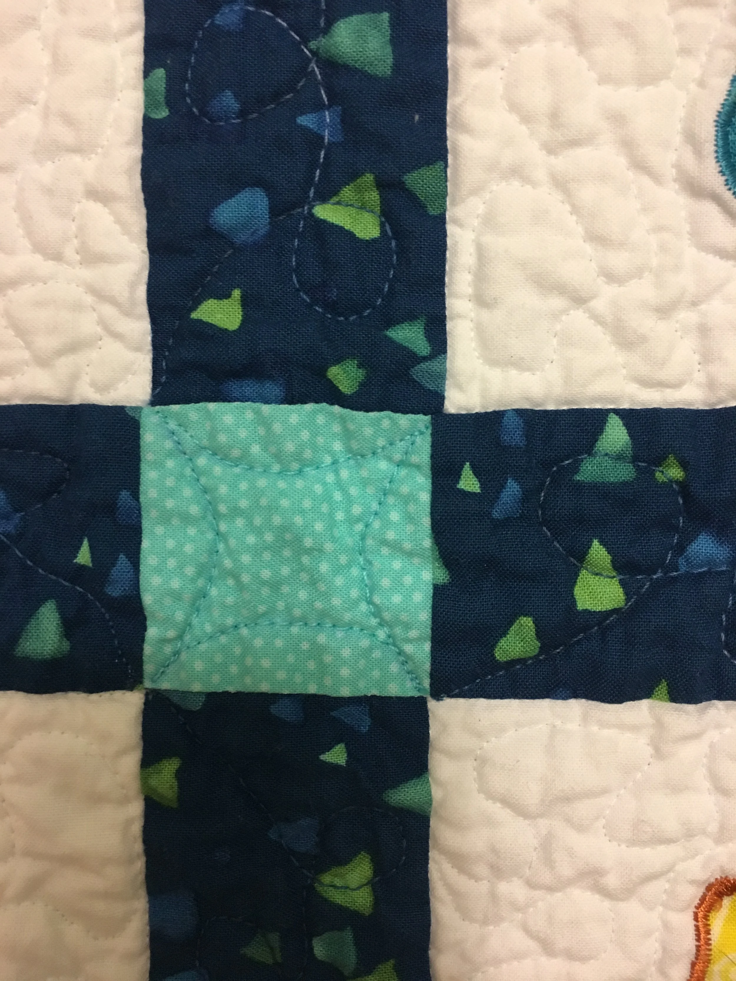 Close-up of a quilt block with a light green square in the center, surrounded by dark blue squares with green and blue accents, and white background pieces, all stitched together.