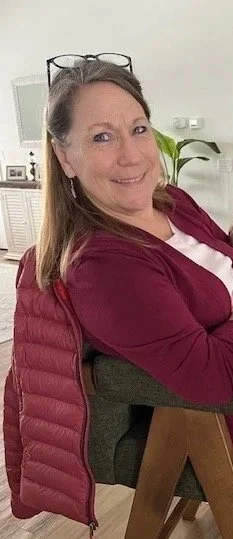 Smiling woman with glasses on her head sitting at a desk in an office or home setting, with a plant and framed pictures in the background.