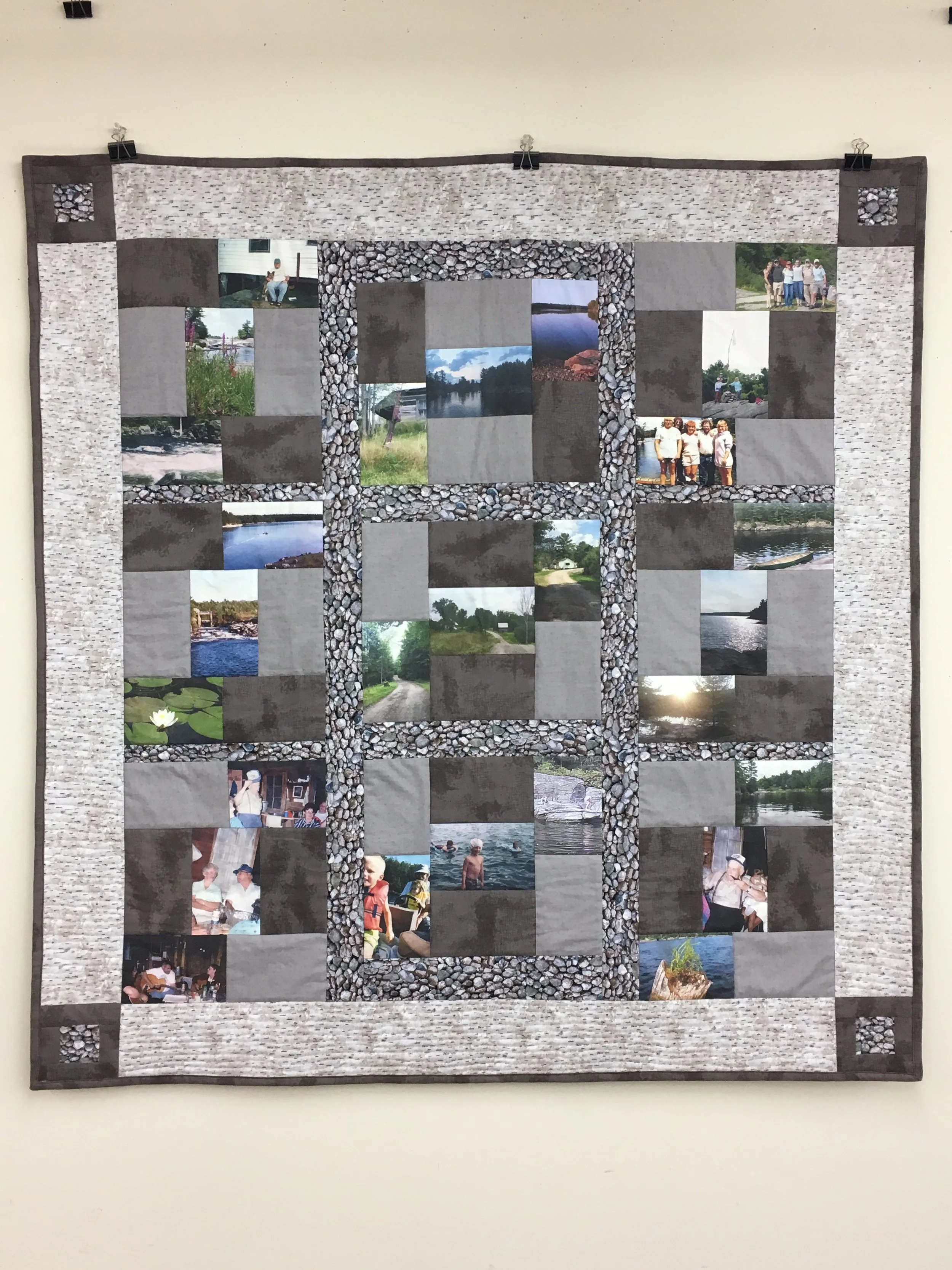 A quilt hanging on a wall with a pattern of photos and fabric squares arranged in a grid, bordered by a pebble-patterned fabric and a light-colored fabric with a textured pattern.