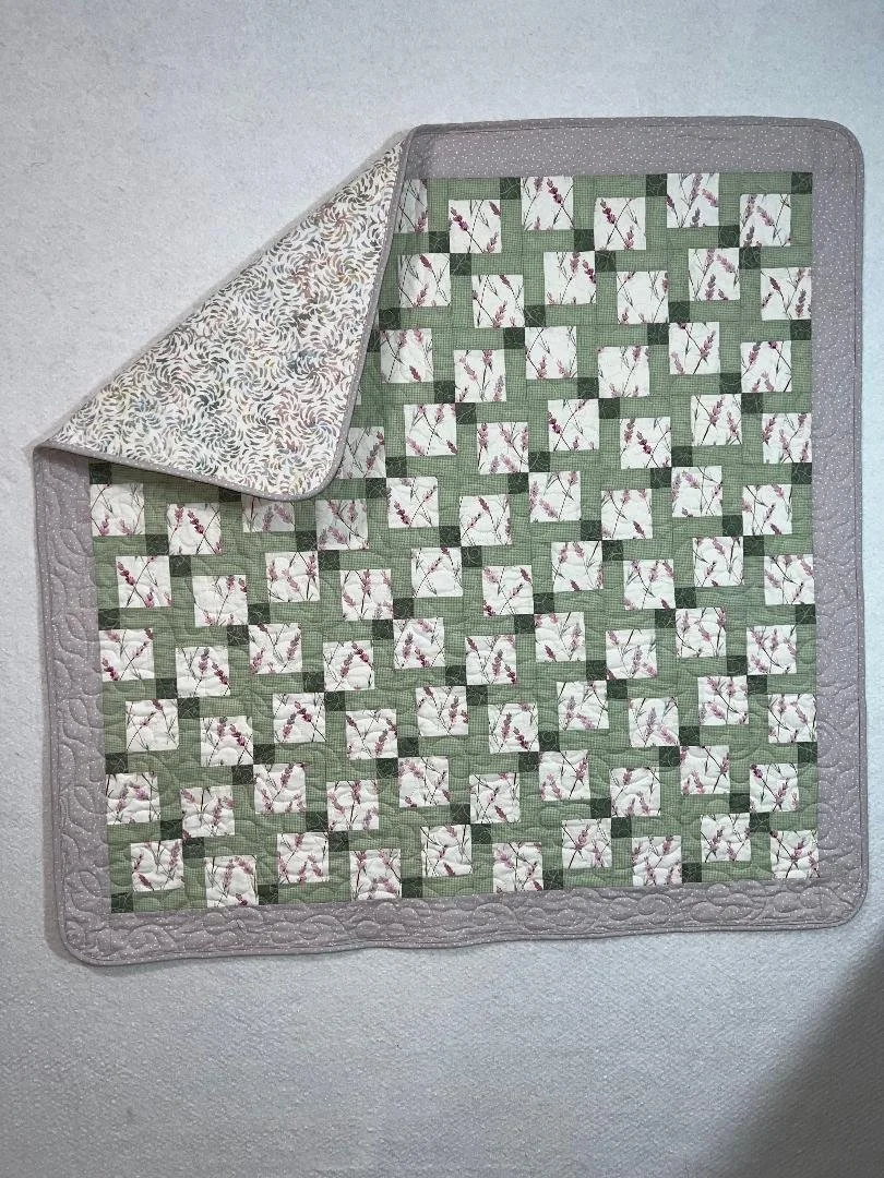 A quilt with a green and white checked pattern and floral designs, bordered with a light gray fabric, draped over a white textured surface.