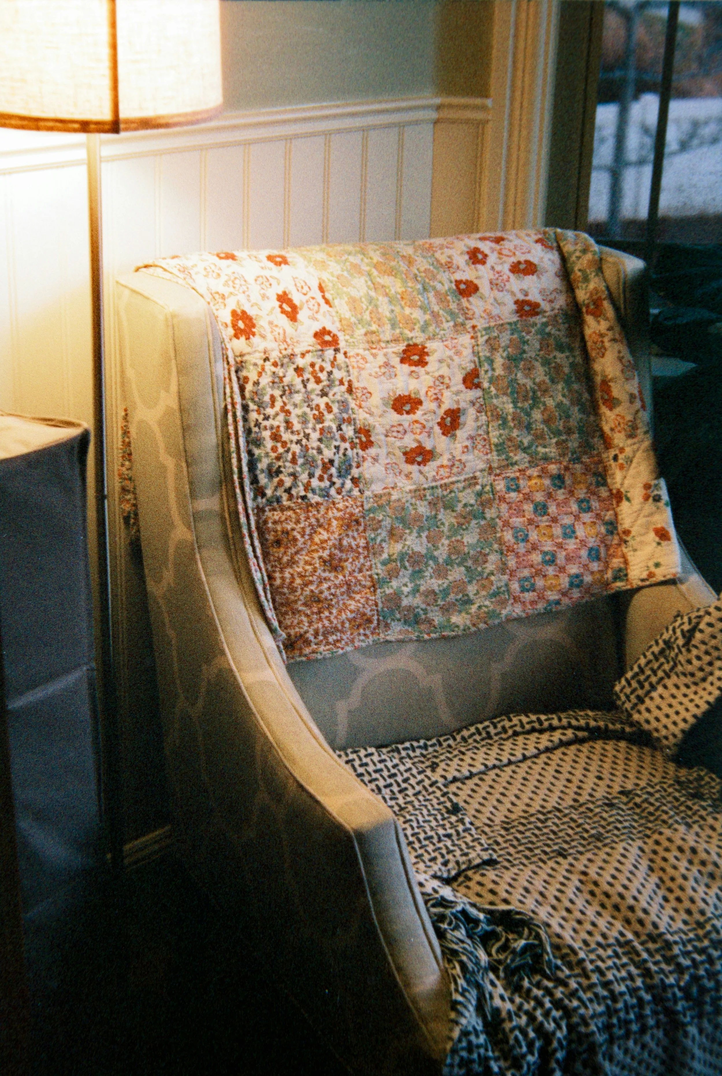 A beige armchair with a colorful patchwork quilt draped over the backrest, next to a tall lamp in a cozy room with a window showing snow outside.