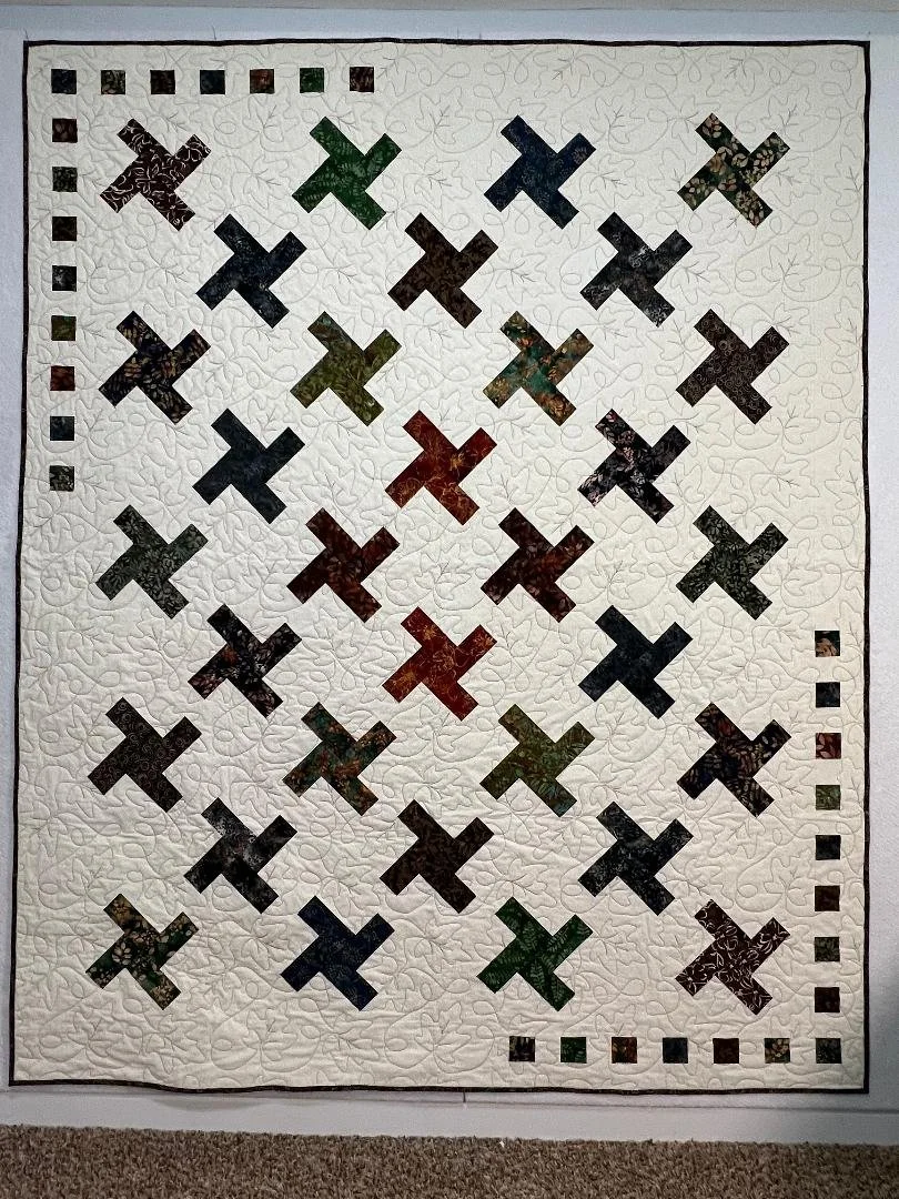 A quilt with a white background and a pattern of various colored X shapes, bordered by smaller square designs in alternating colors.
