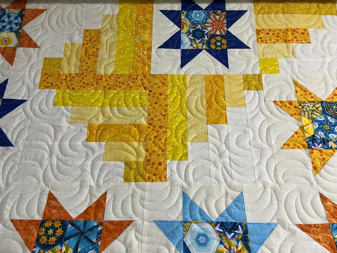 Close-up of a quilt with stars and crosses in yellow, orange, blue, and white fabric with intricate stitching details.