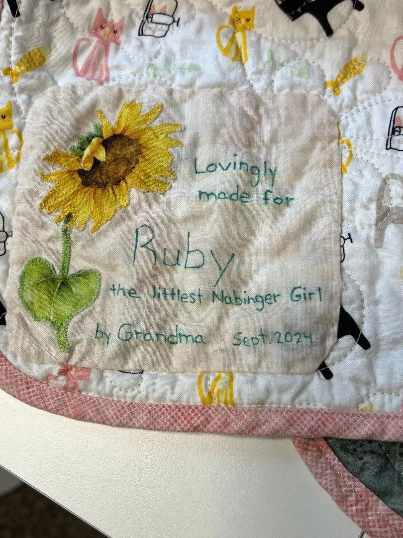 A fabric cloth with embroidered text and a sunflower design. The text reads: "Lovingly made for Ruby the littlest Nabinger Girl by Grandma Sept. 2024." The surrounding quilt features colorful cats and other whimsical patterns.