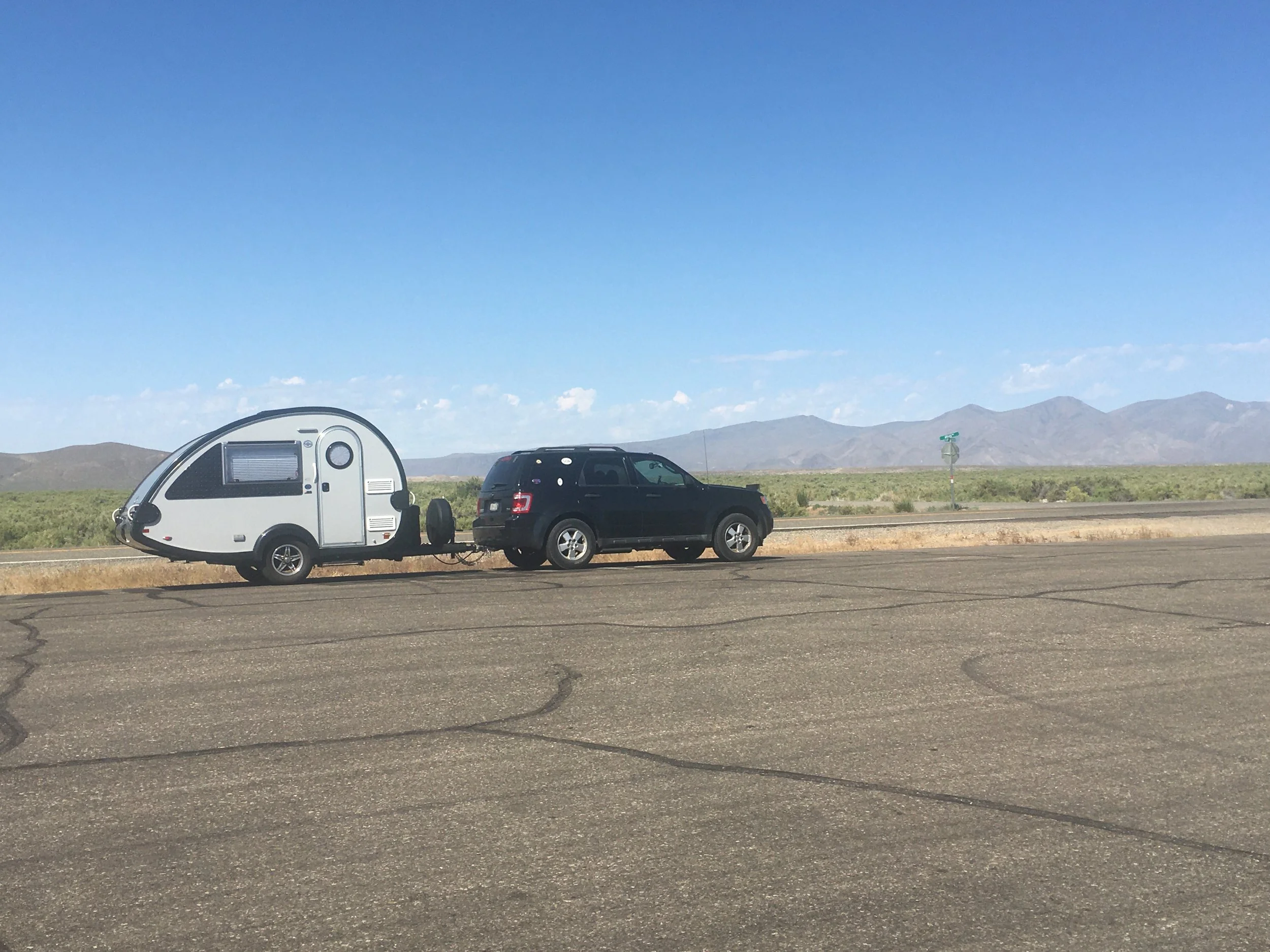 A black SUV towing a small travel trailer with a rounded shape, parked on the side of a wide, empty paved road with a desert landscape and mountains in the background, under a clear blue sky.