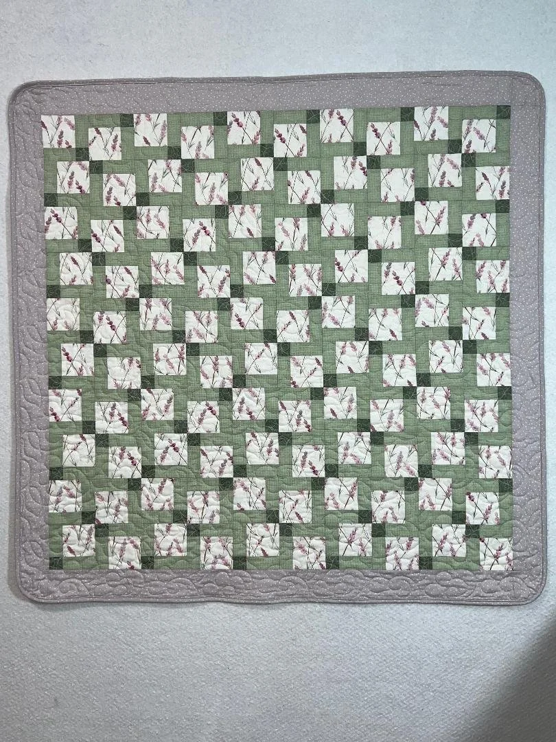 A quilt with a green and white geometric pattern, featuring small square blocks with floral prints, bordered by light gray fabric with a subtle pattern.